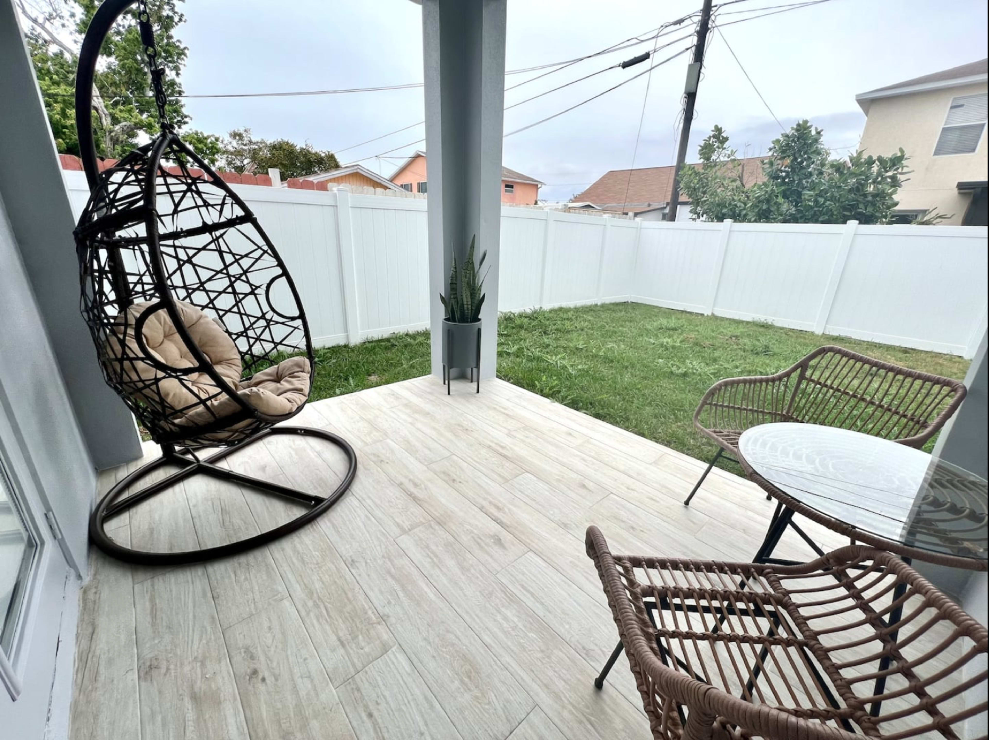 A porch features a hanging chair, a small table, and a view of a fenced yard.
