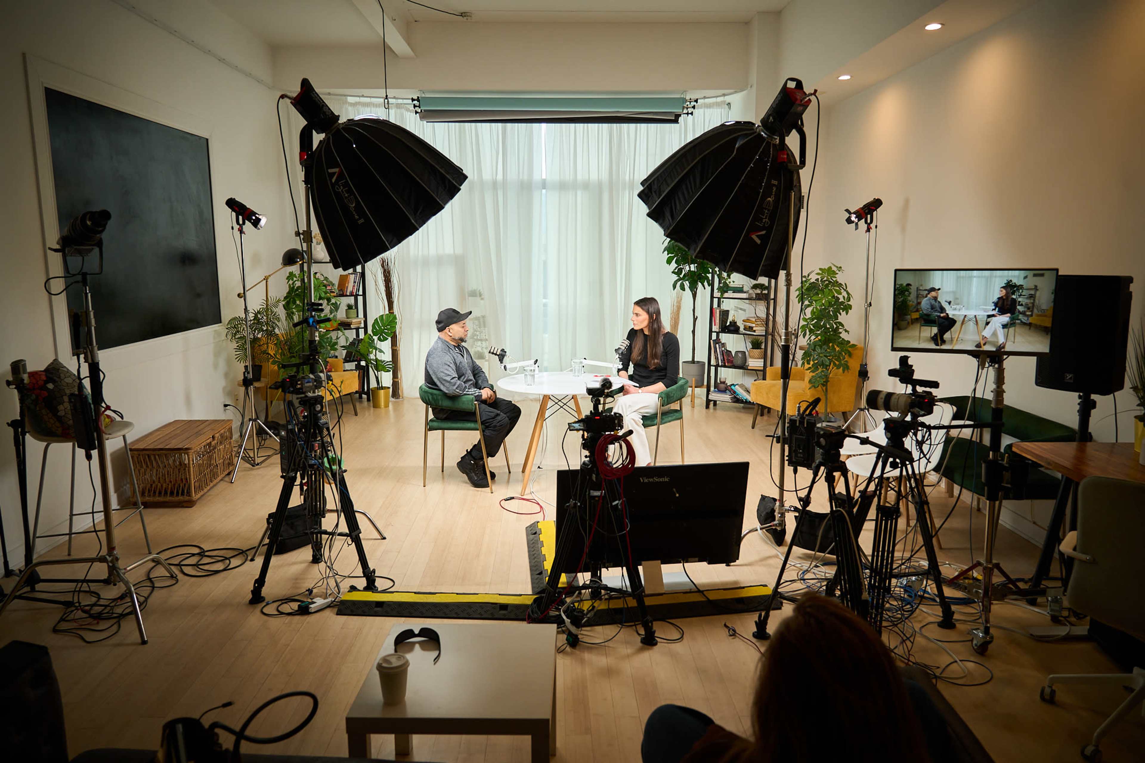 A woman interviews a man in a well-lit studio filled with cameras, lights, and plants.