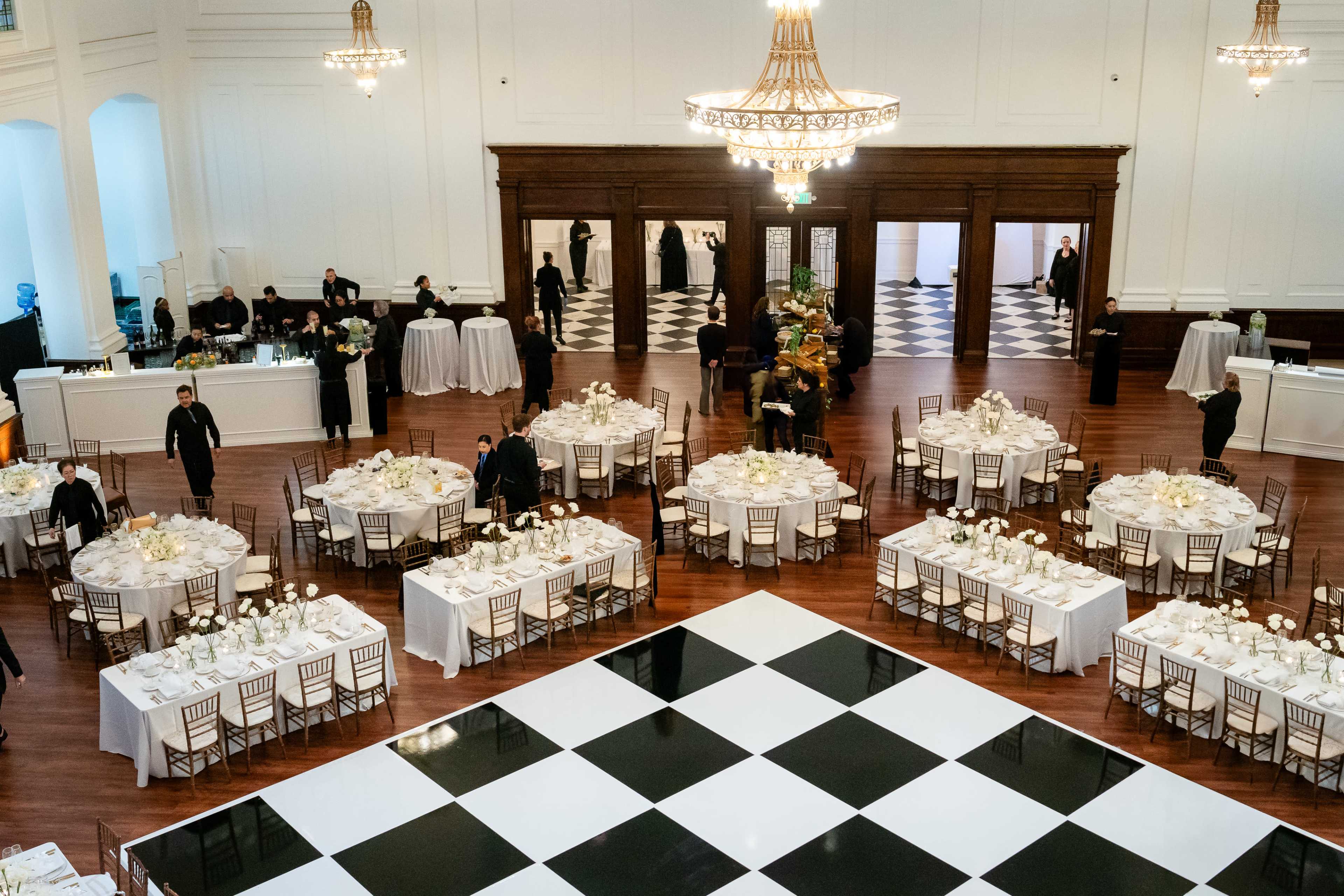 The image shows a large banquet hall set for an event, featuring several tables arranged with white tablecloths, centerpieces, and a black and white checkered dance floor in the center.