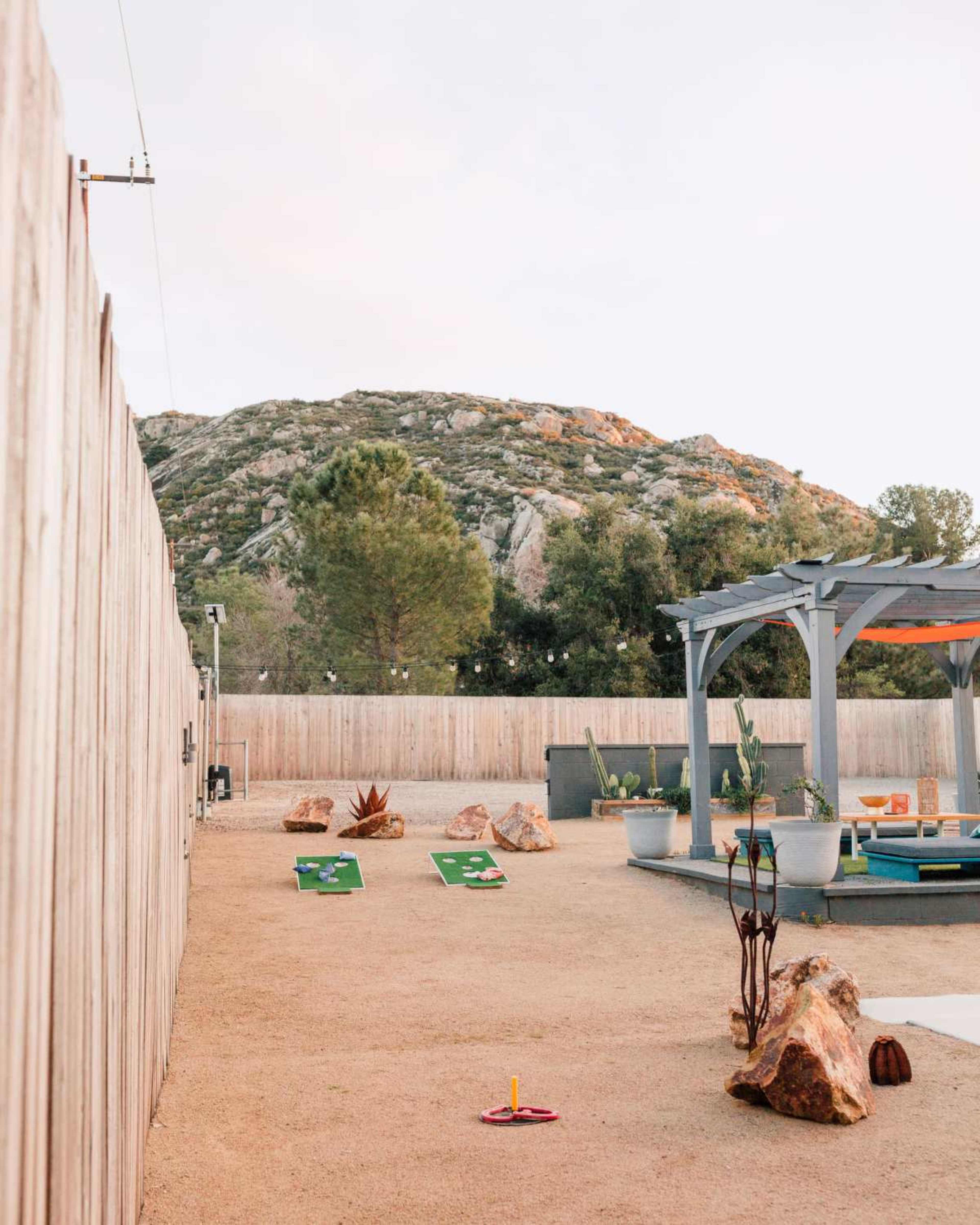 A landscaped outdoor area features a mini-golf setup with green mats and rocks, framed by a wooden fence and backed by a rocky hillside.