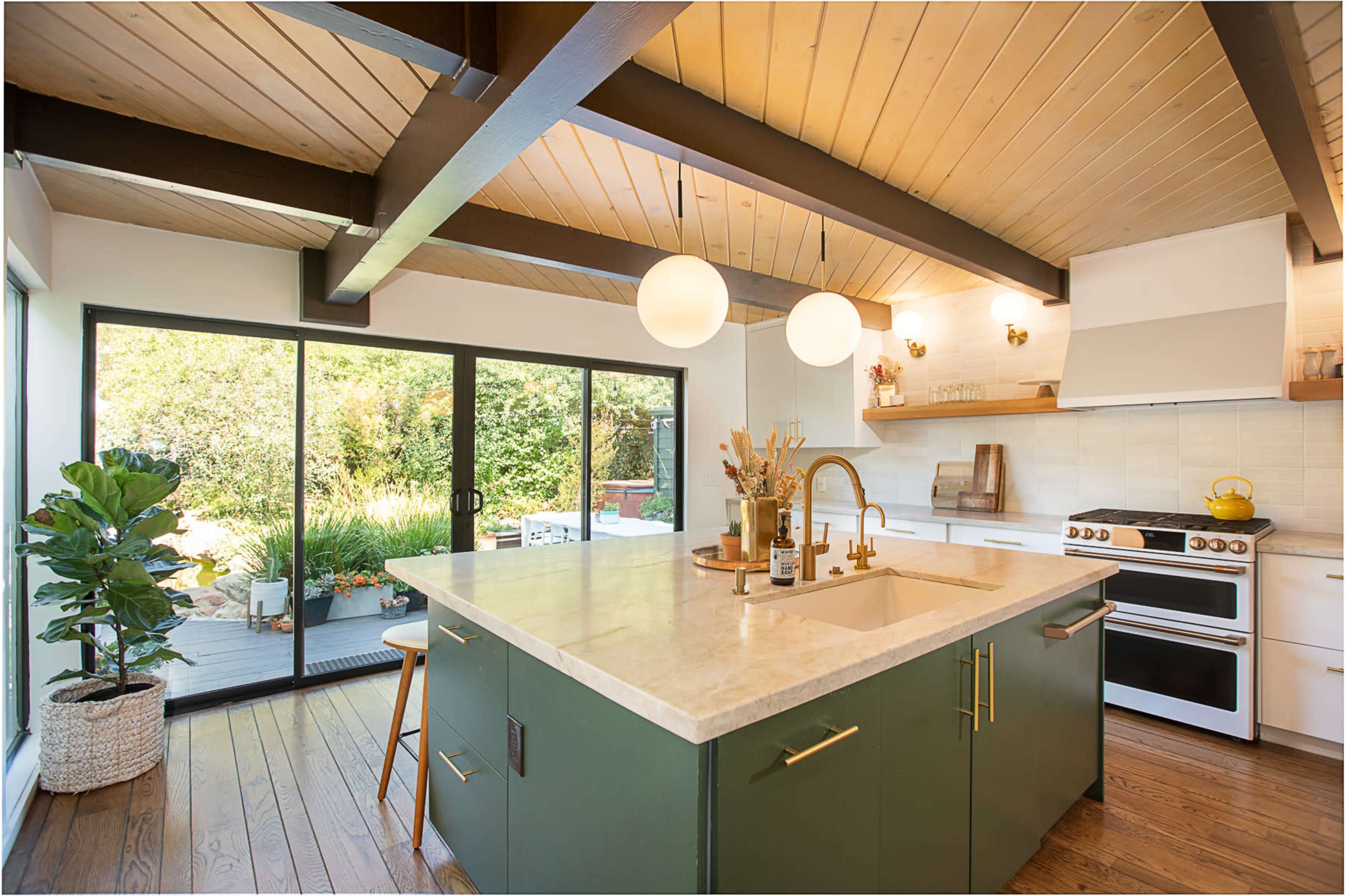A modern kitchen features a central island with a light-colored countertop, green cabinetry, and large windows overlooking a garden.
