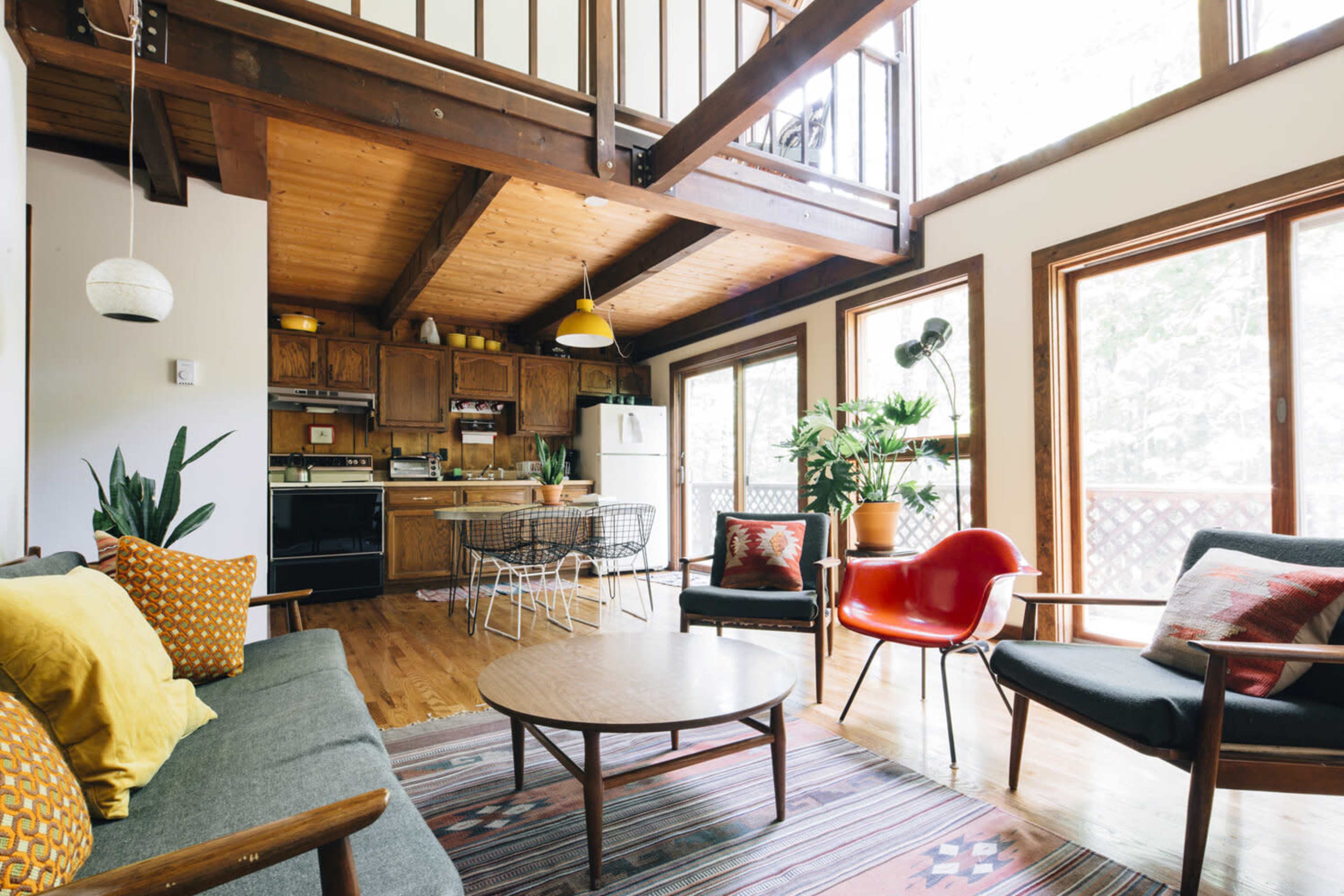 The image shows a spacious living area with wooden beams, a modern kitchen in the background, and large windows allowing natural light to enter.