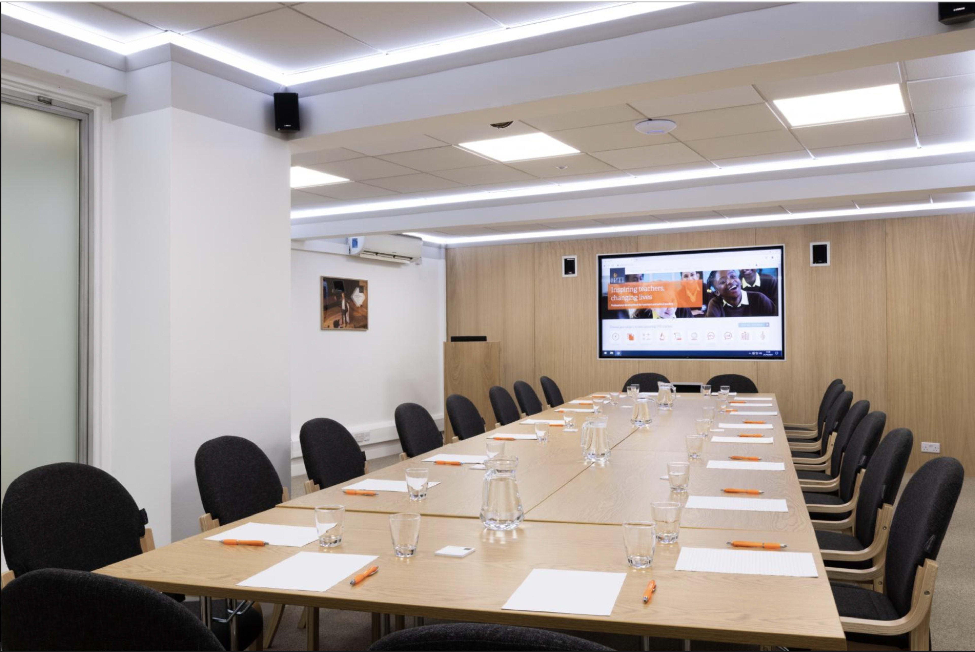 A modern conference room features a long wooden table set with notebooks and pens, facing a large screen displaying digital content.