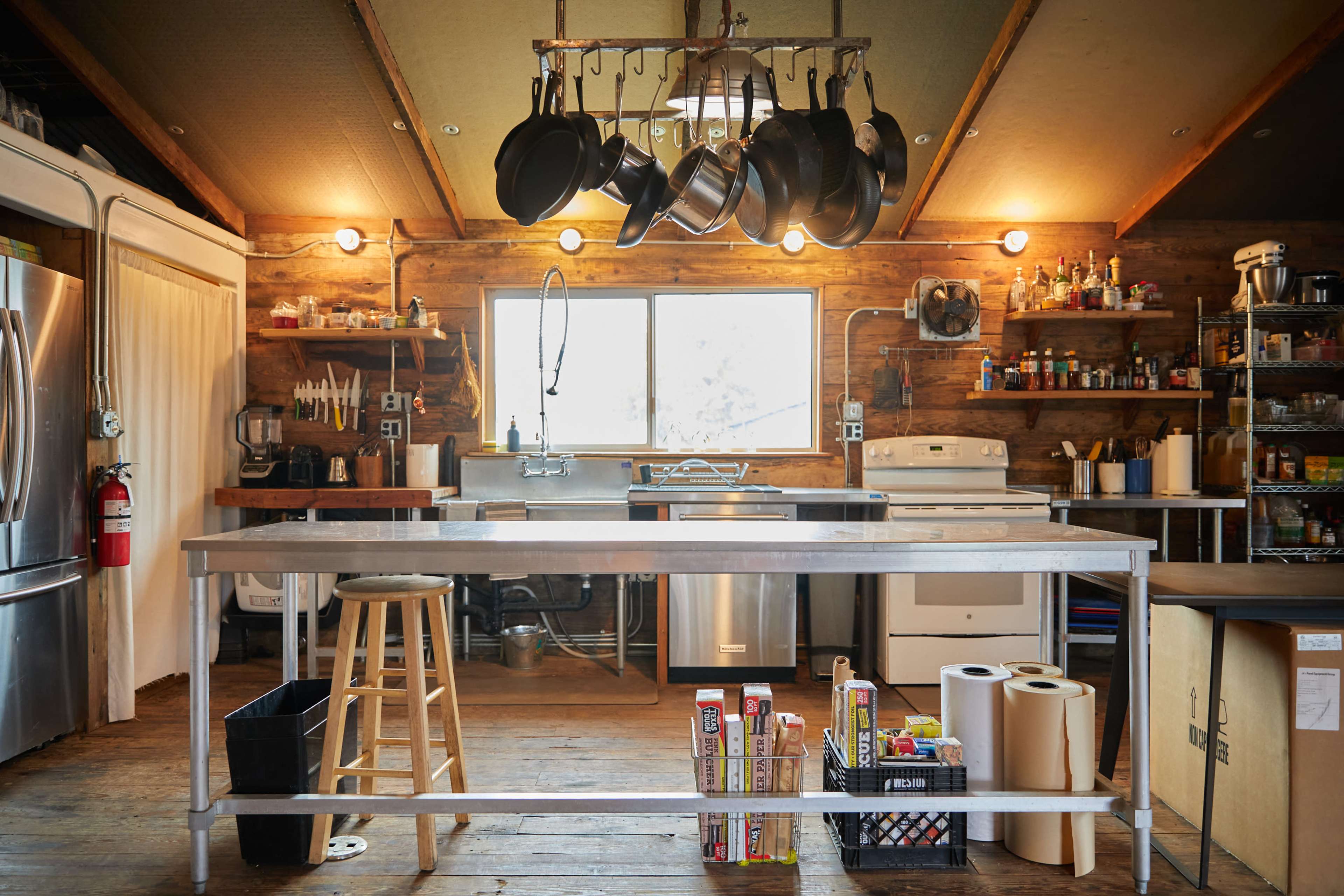 A rustic kitchen with a central metal island, hanging pots above, and various appliances and shelves along the walls.
