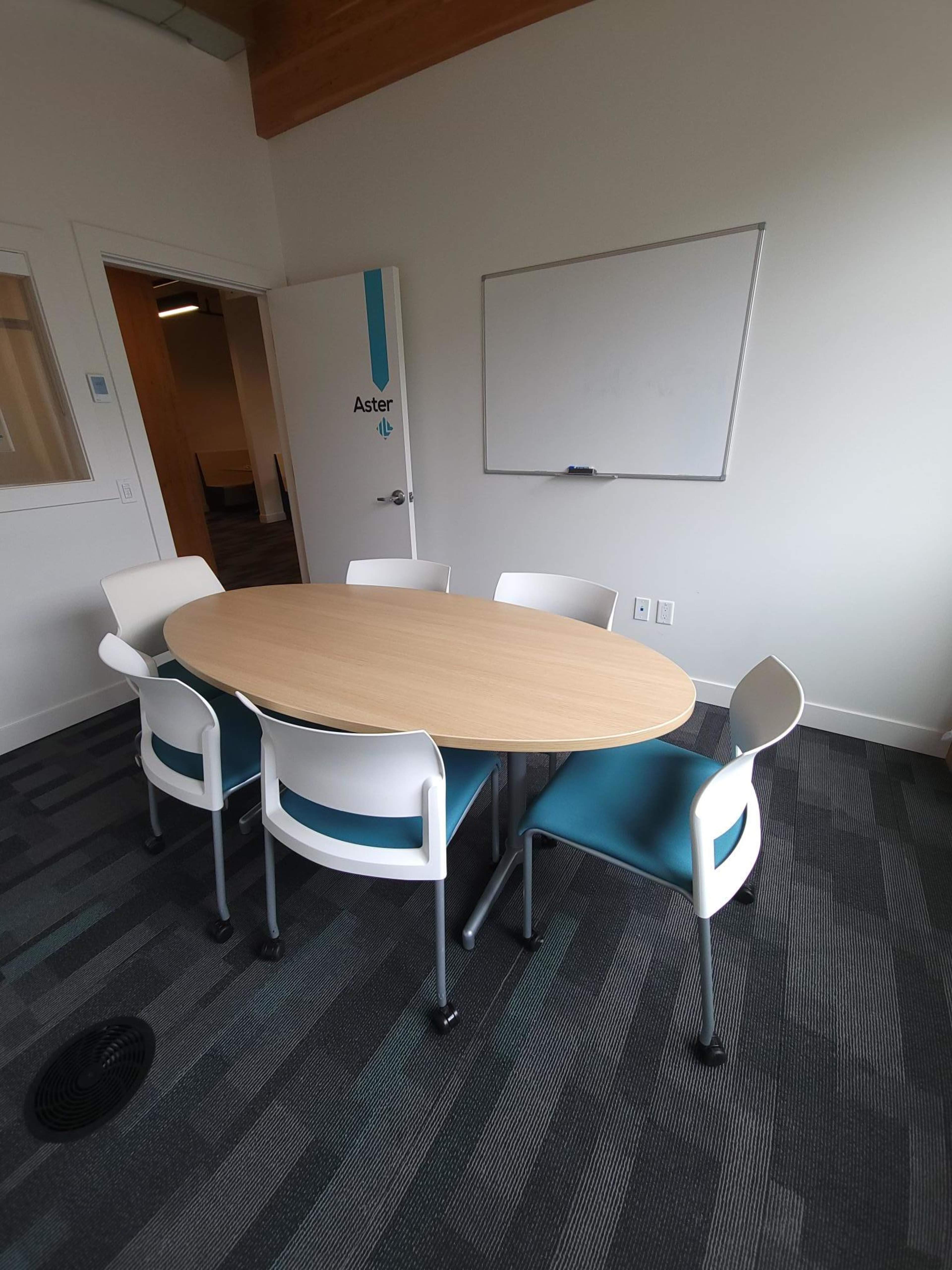A round wooden table surrounded by five white and blue chairs in a conference room with a whiteboard and a door labeled "Aster."