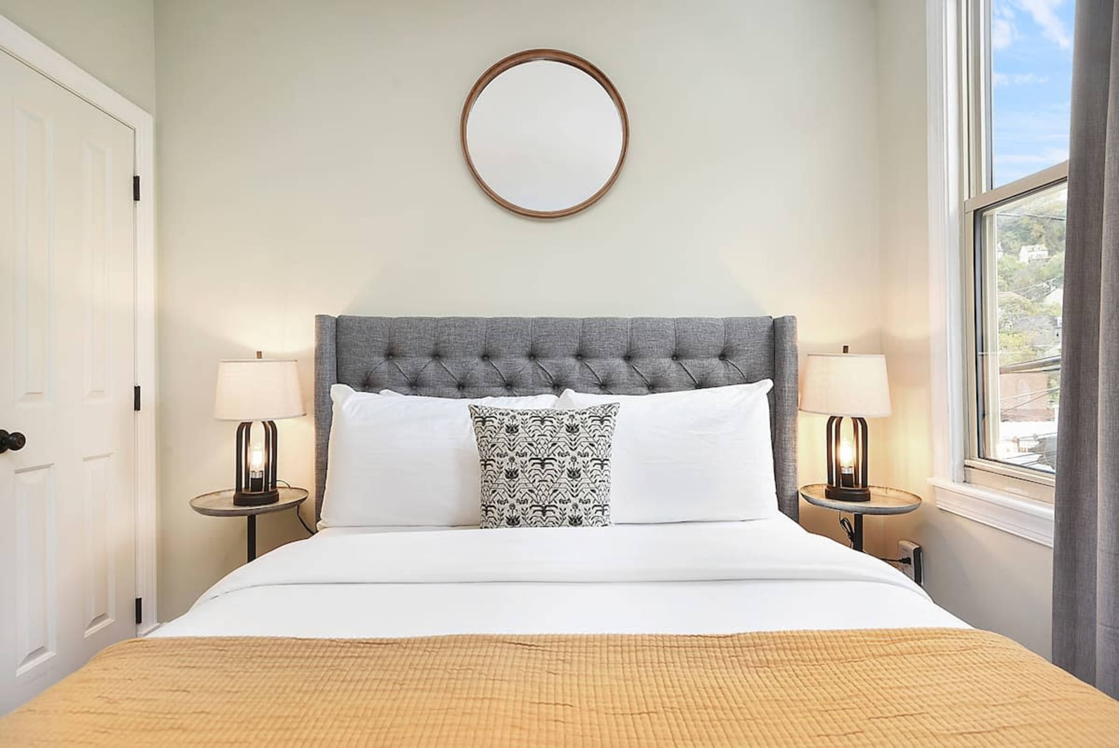 The image shows a neatly made bed with a gray upholstered headboard, two bedside tables with lamps, and a round mirror above the headboard in a light-colored room.