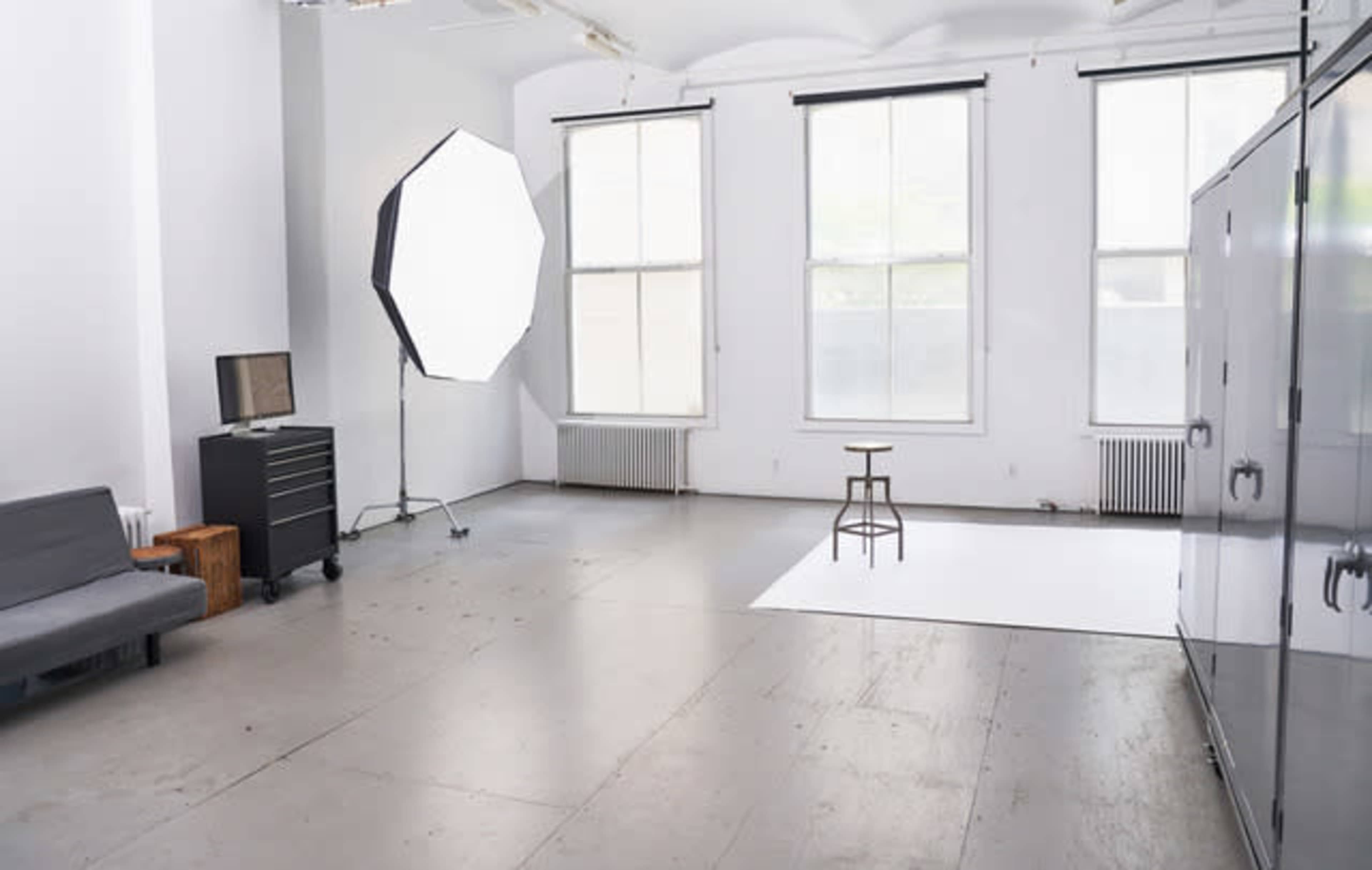 The image shows a spacious, well-lit photography studio featuring a large softbox, a single stool on a white backdrop, and a gray couch against the wall.