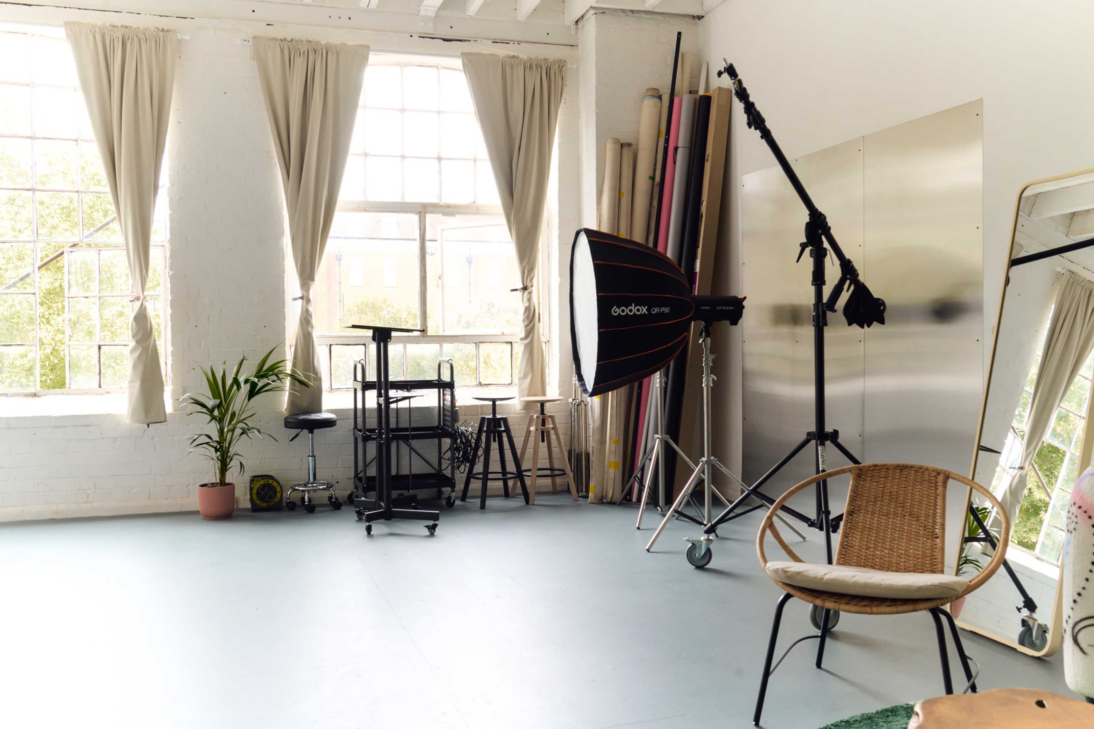The image shows a well-lit photography studio featuring a large softbox light, a rolling cart, a wooden chair, and various colored backdrops.