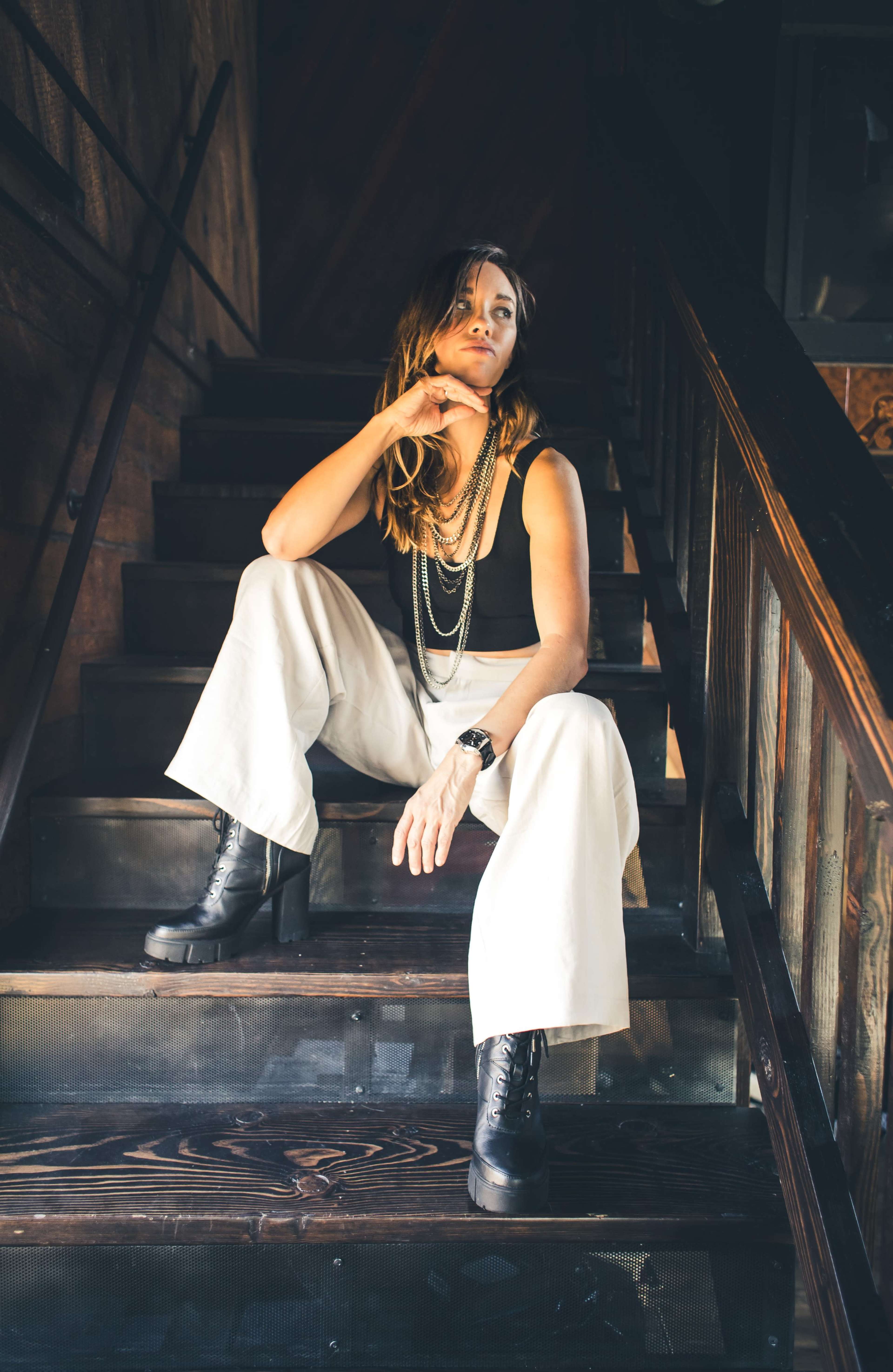 A woman sits on a staircase, wearing a black top and white wide-leg pants, with her chin resting on her hand.