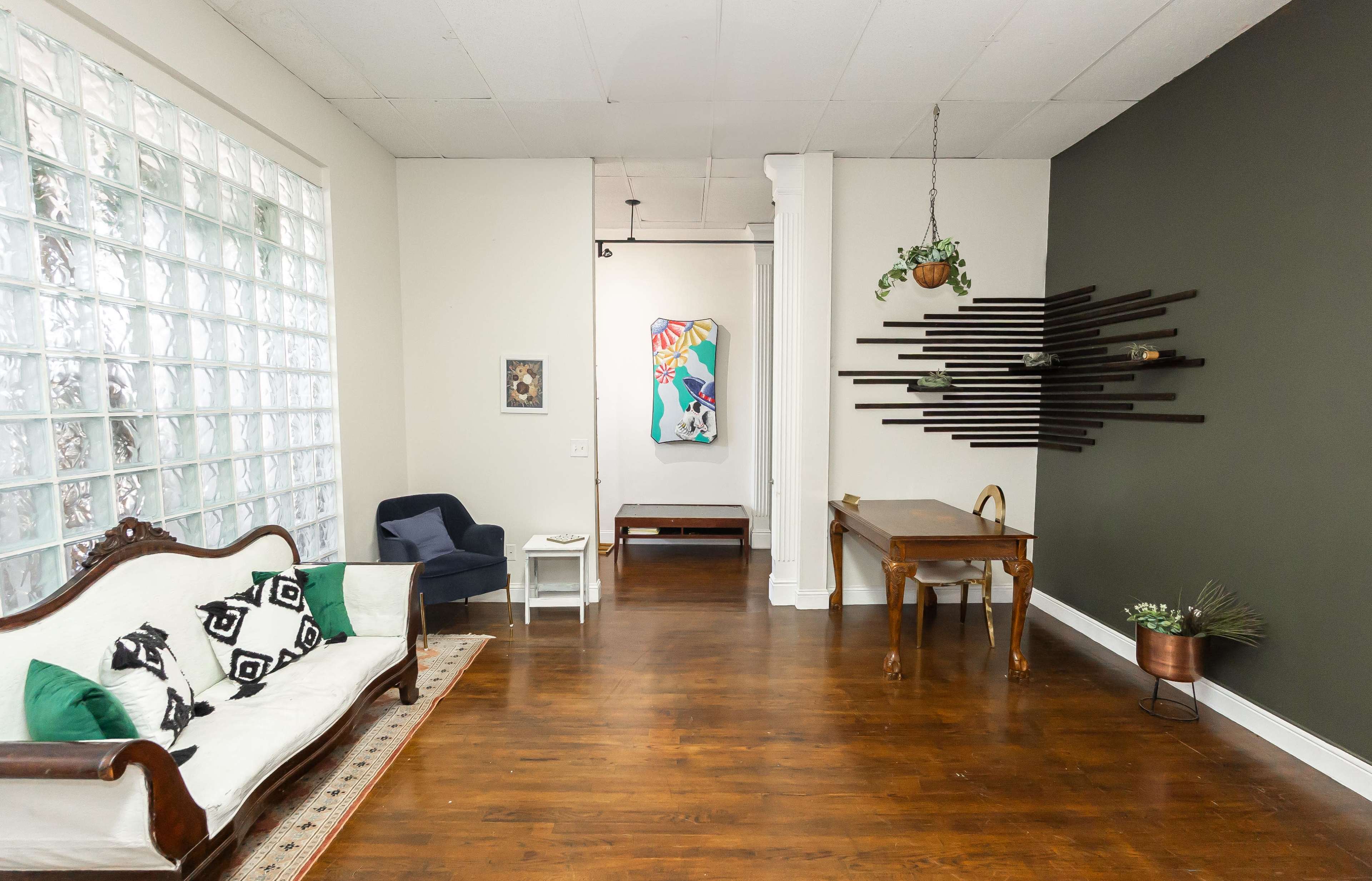 The scene shows a spacious room with a glass block wall on one side, a vintage sofa, a blue armchair, and a wooden dining table against a dark green wall, with artwork displayed in the background.