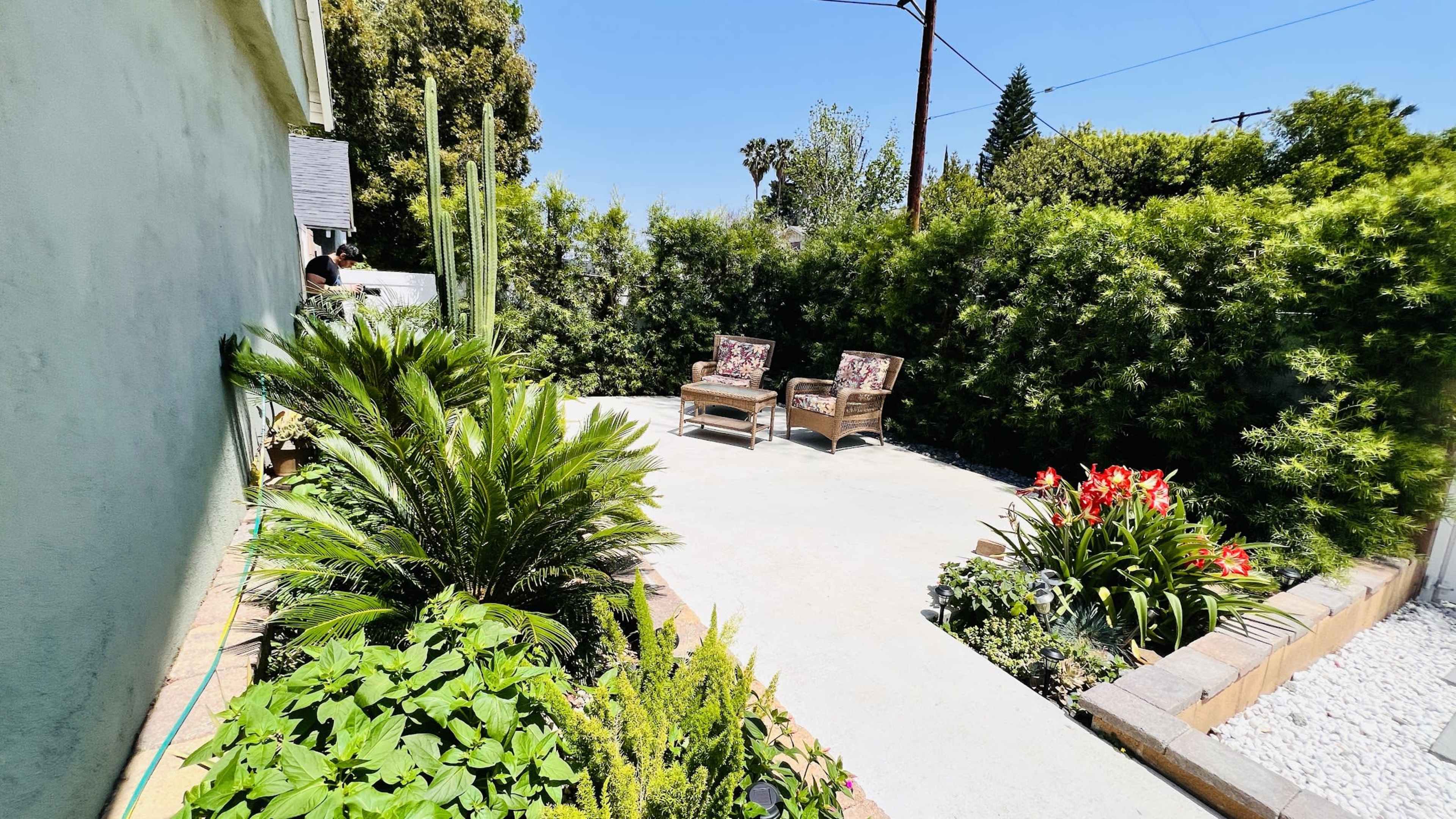 The image shows a small outdoor patio area with two wicker chairs, surrounded by greenery and flowering plants.