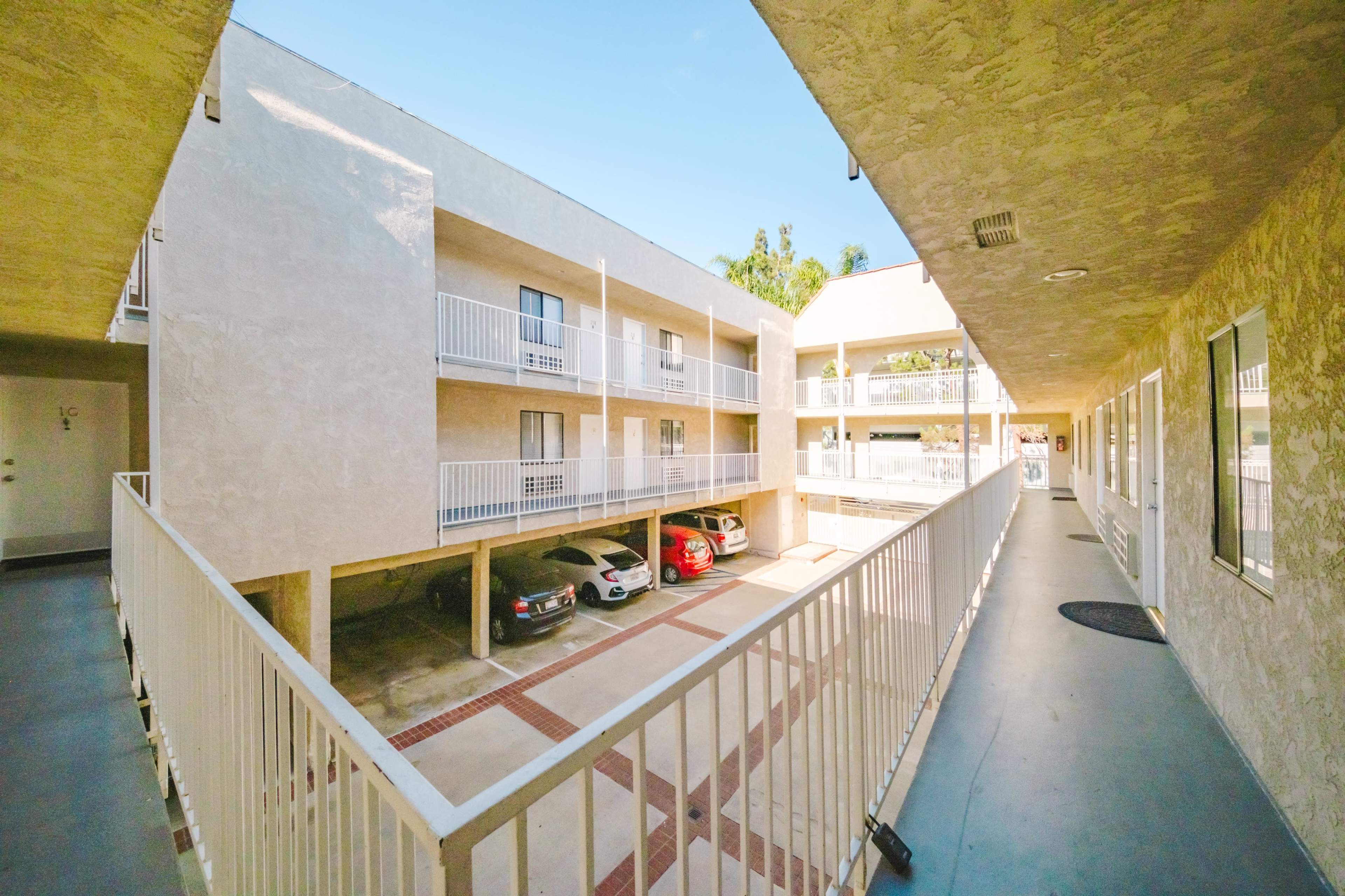 The image shows a courtyard view of a multi-level apartment building with cars parked underneath and balconies overlooking the open space.