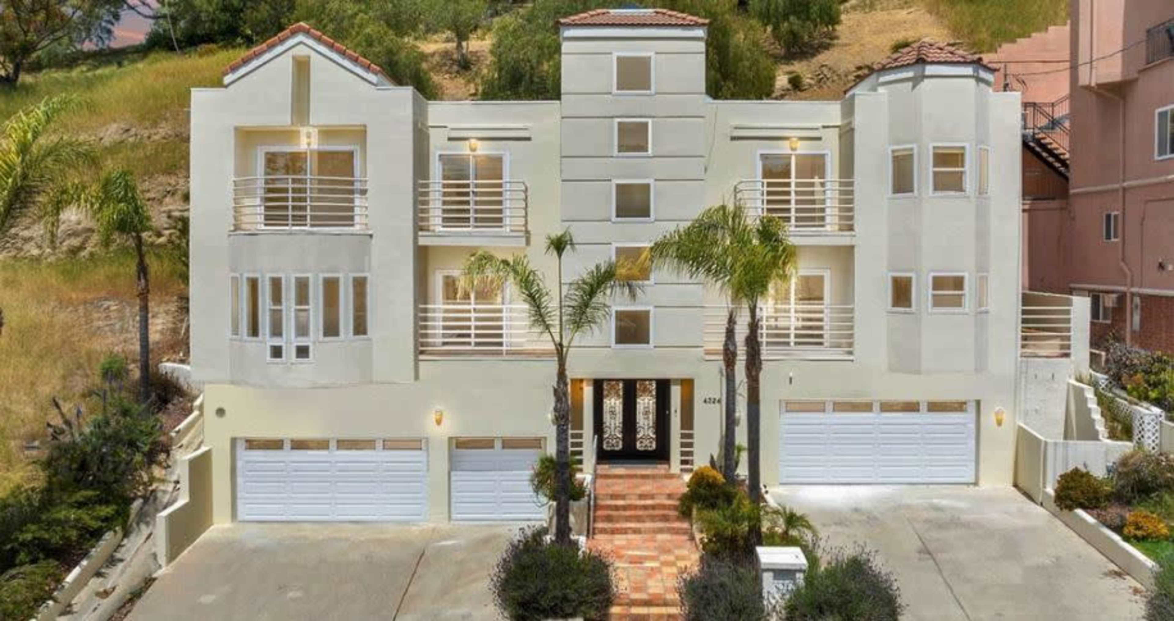 A three-story modern home with multiple balconies, a driveway with three garage doors, and landscaping featuring palm trees.