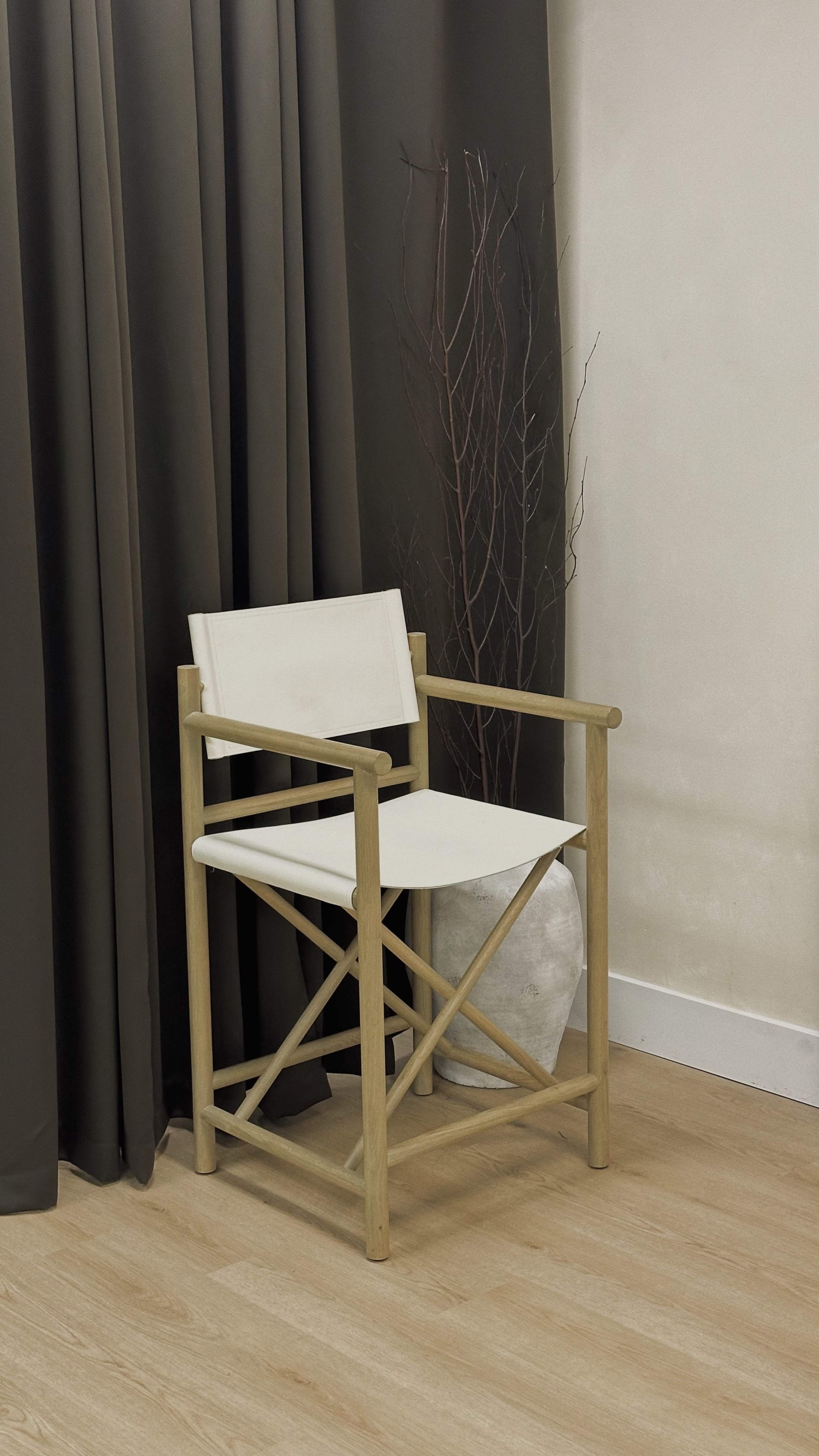 A light-colored wooden chair with a white fabric seat is positioned against a backdrop of dark curtains and a decorative branch.