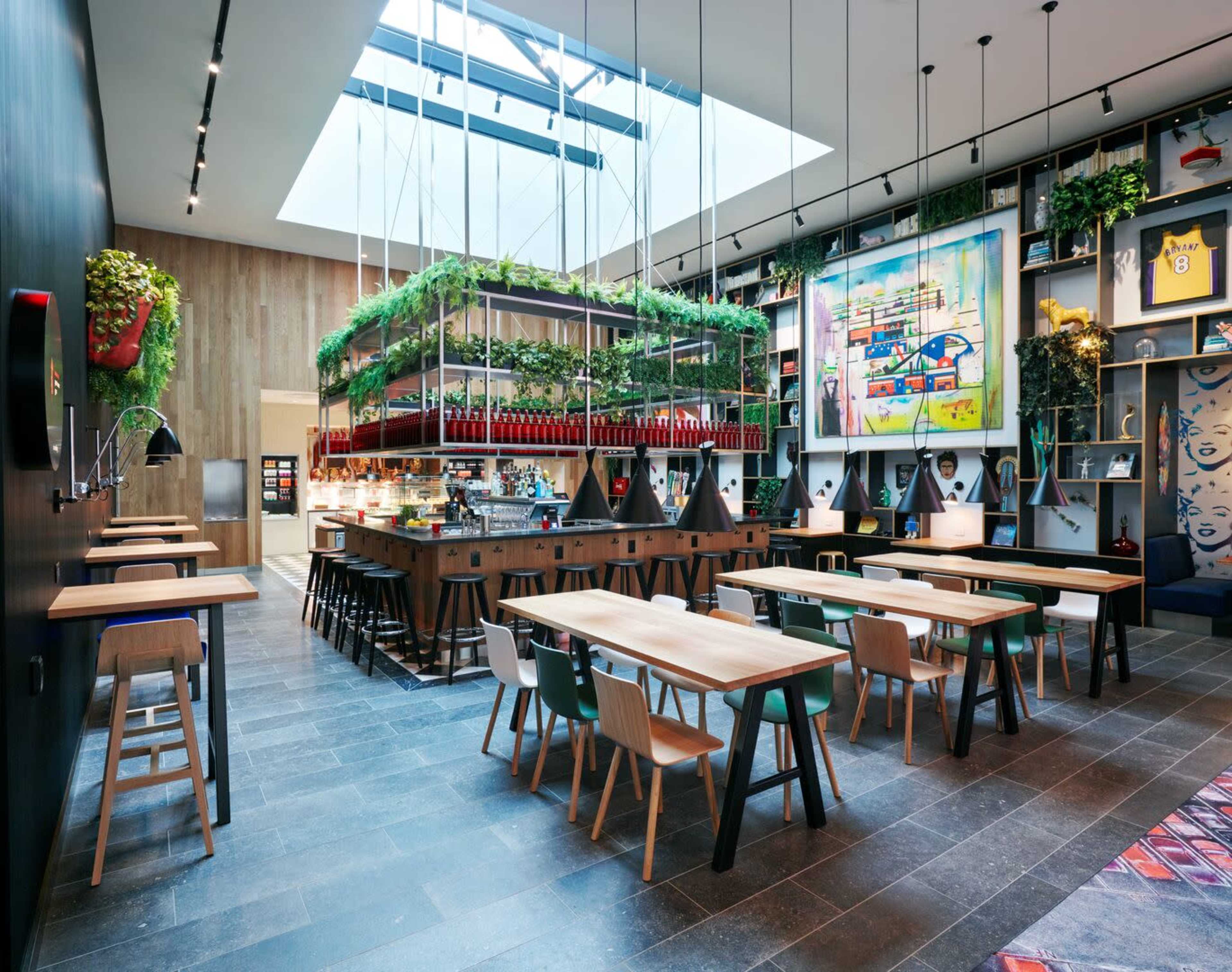 The image shows a modern café interior featuring wooden tables, a hanging garden above the counter, and large skylights illuminating the space.