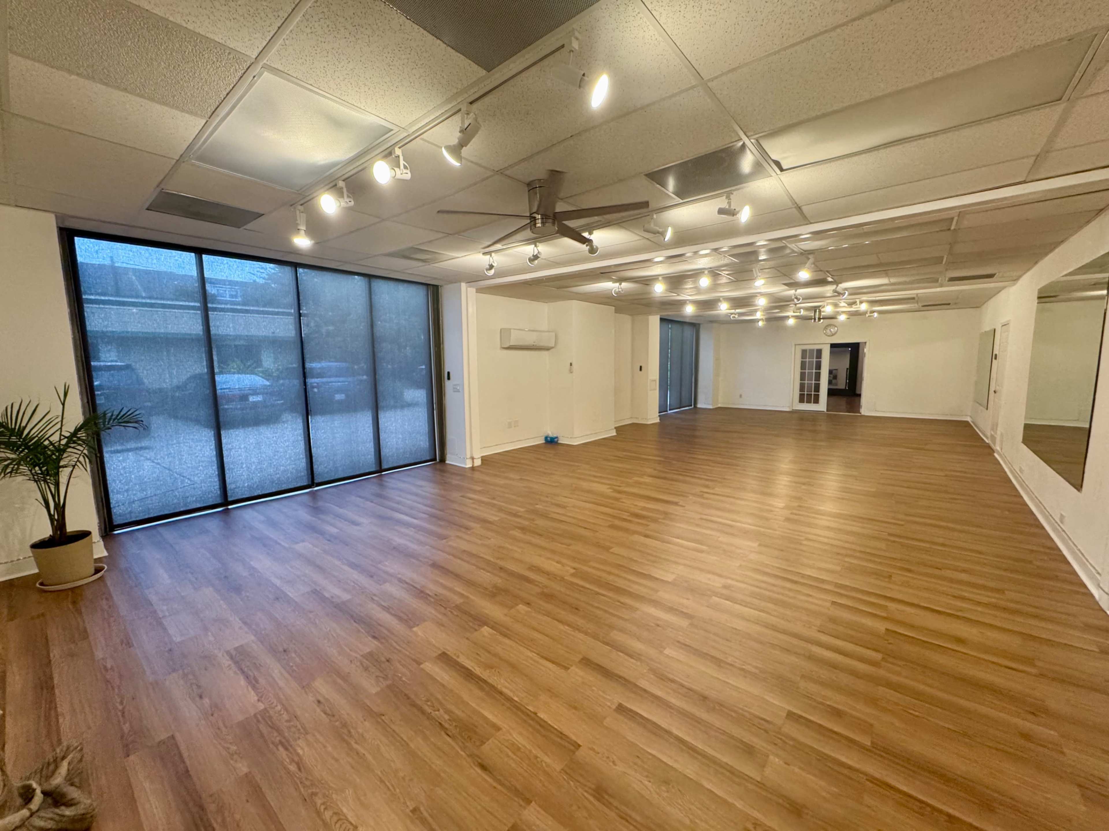The image shows an empty room with wooden flooring, large windows, and overhead lighting.