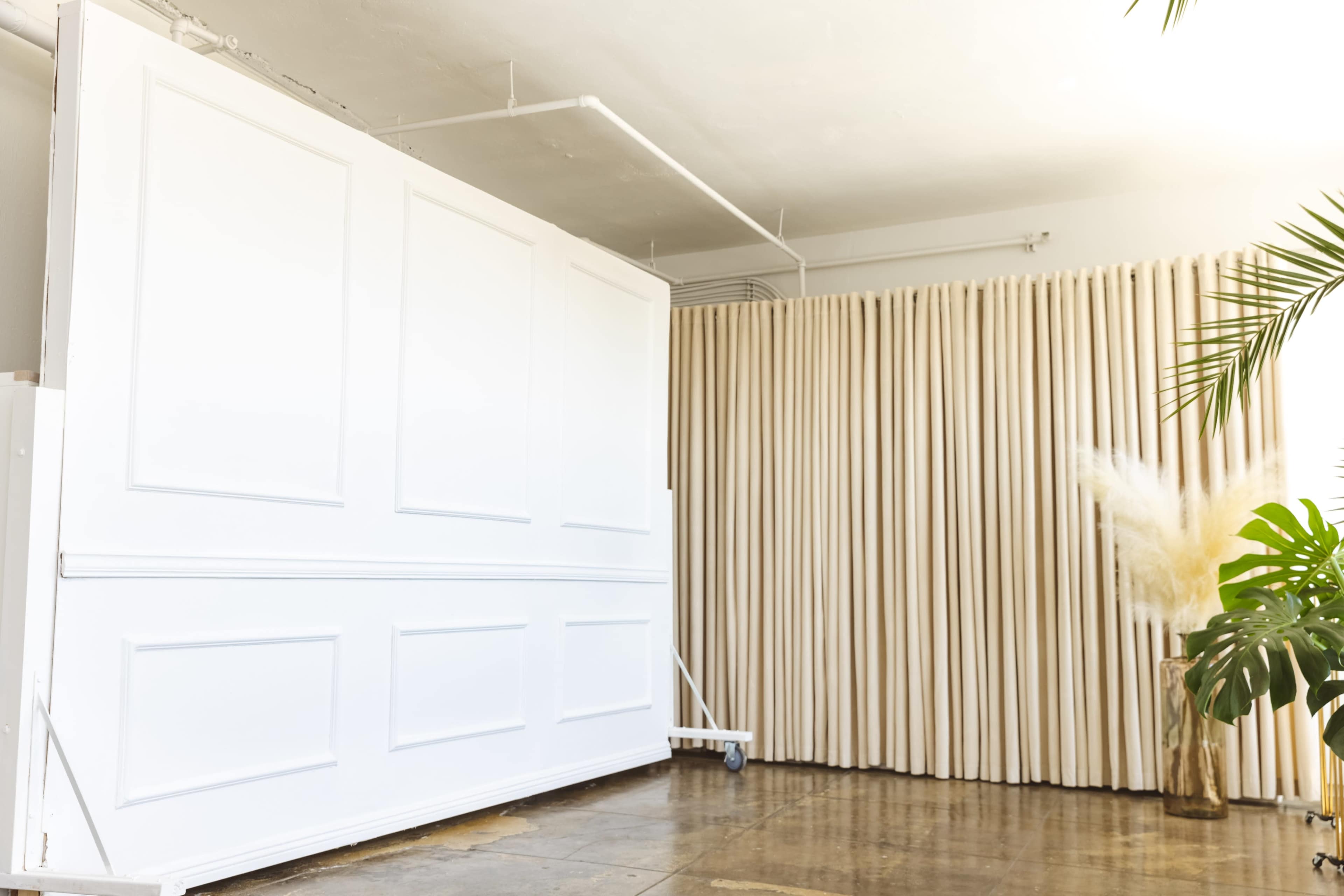 The image shows a spacious interior with a large white wall on the left and a curtain of beige fabric on the right, accompanied by decorative plants.