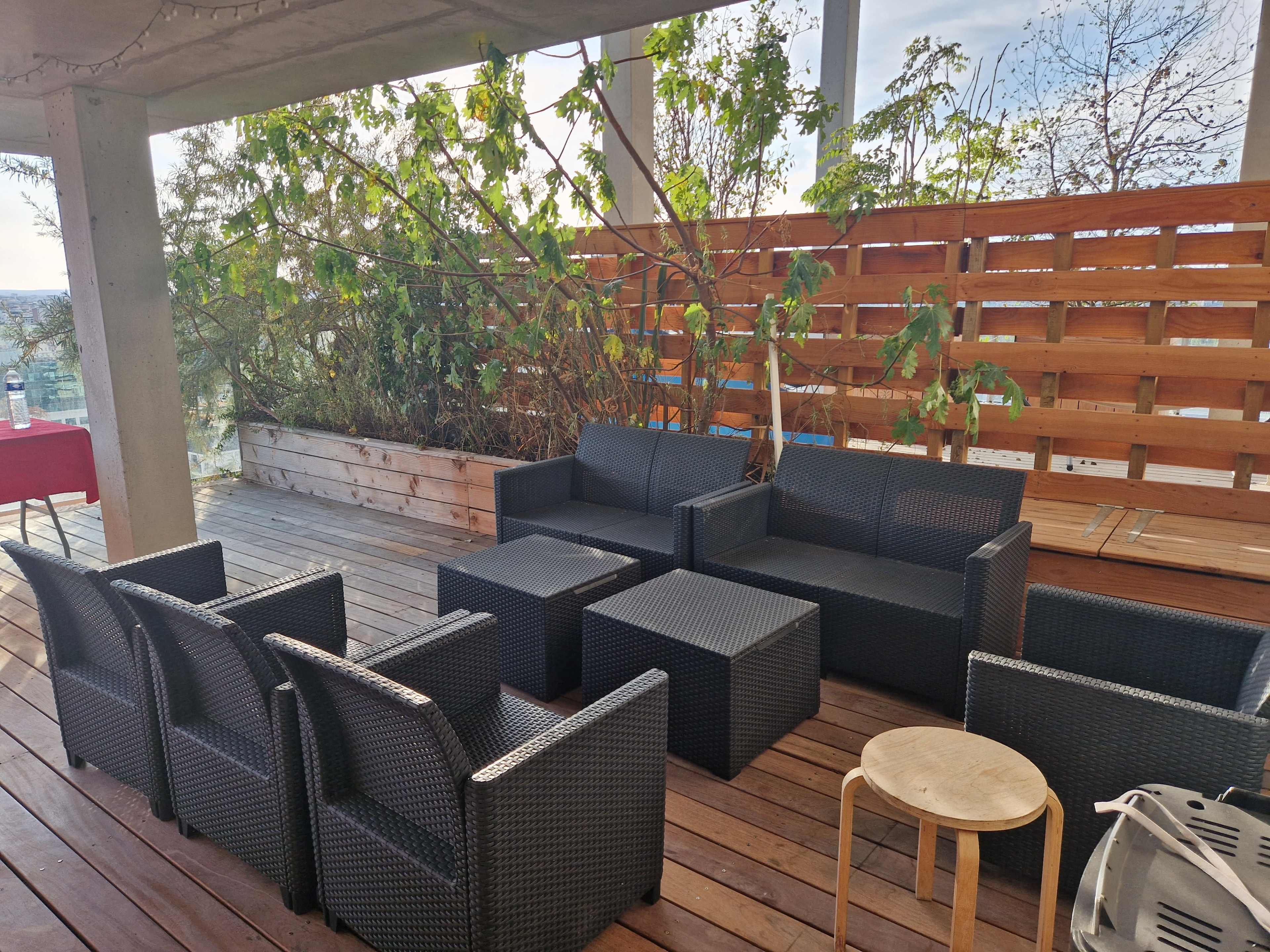 A modern outdoor seating area features a set of black wicker furniture grouped around a low table, surrounded by wooden decking and greenery.