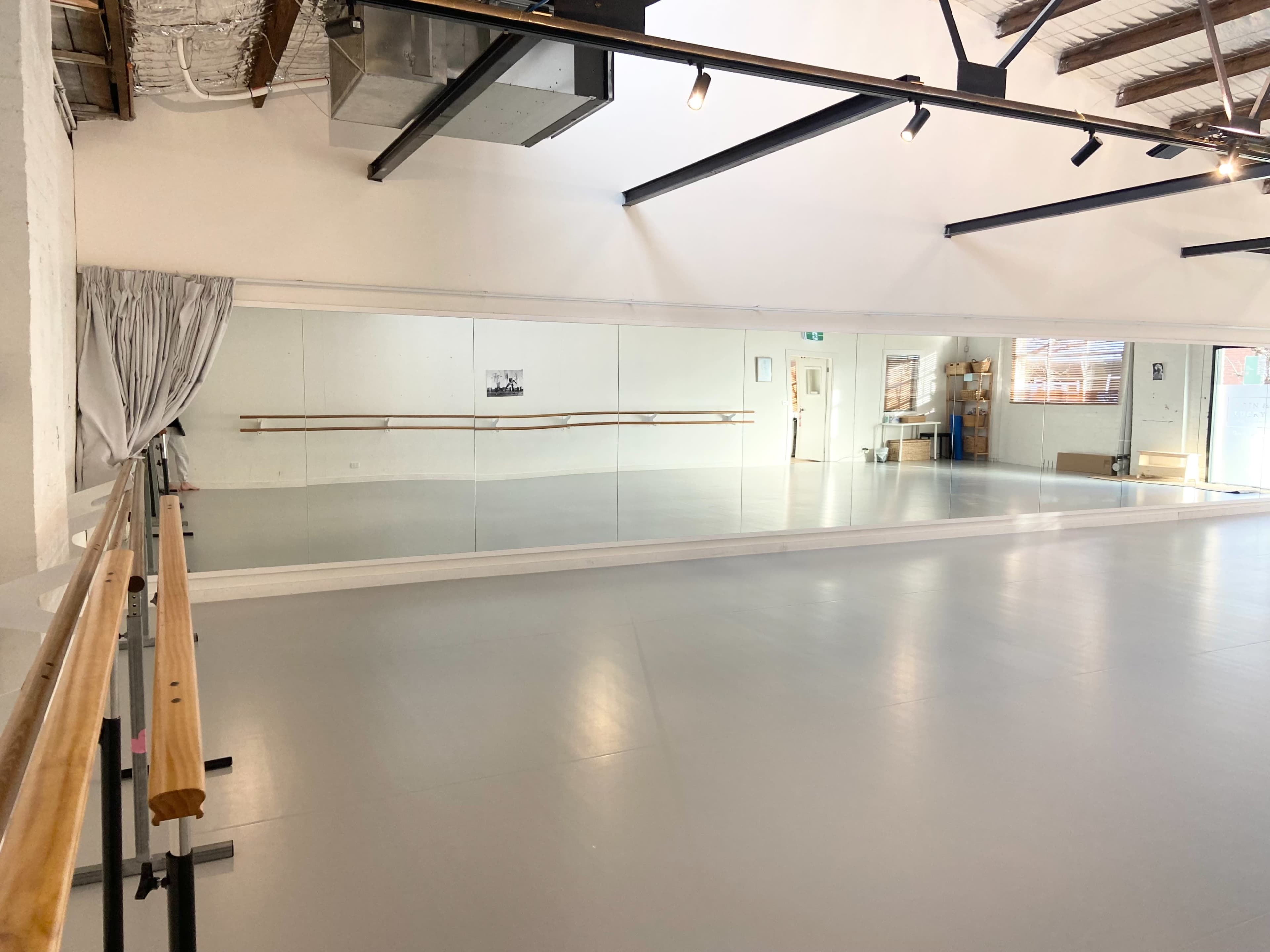 The image shows a spacious ballet studio with a mirrored wall, wooden ballet barre, and a gray floor.