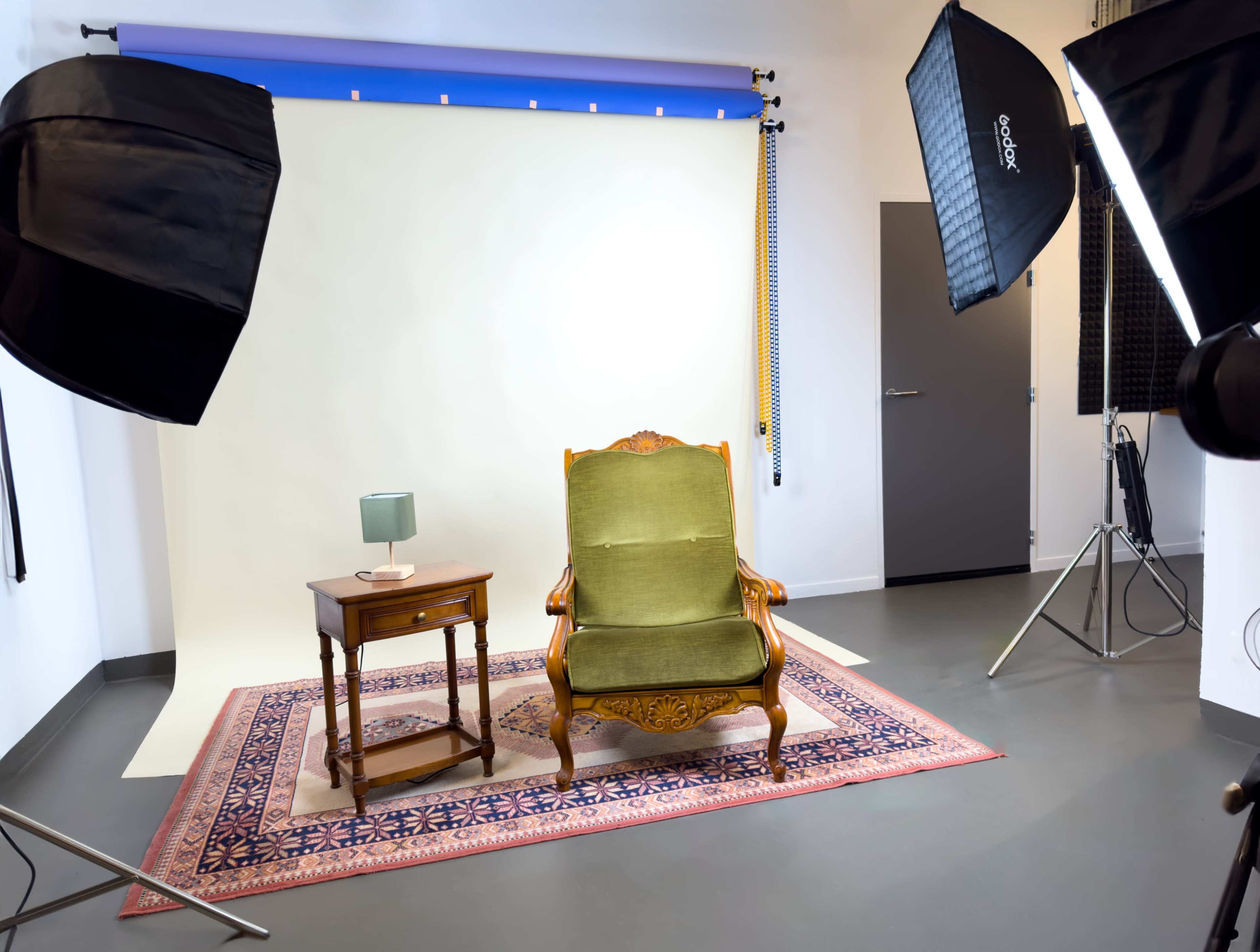 The image shows a green upholstered armchair and a small wooden side table with a lamp, positioned on a patterned rug in a well-lit studio setting.
