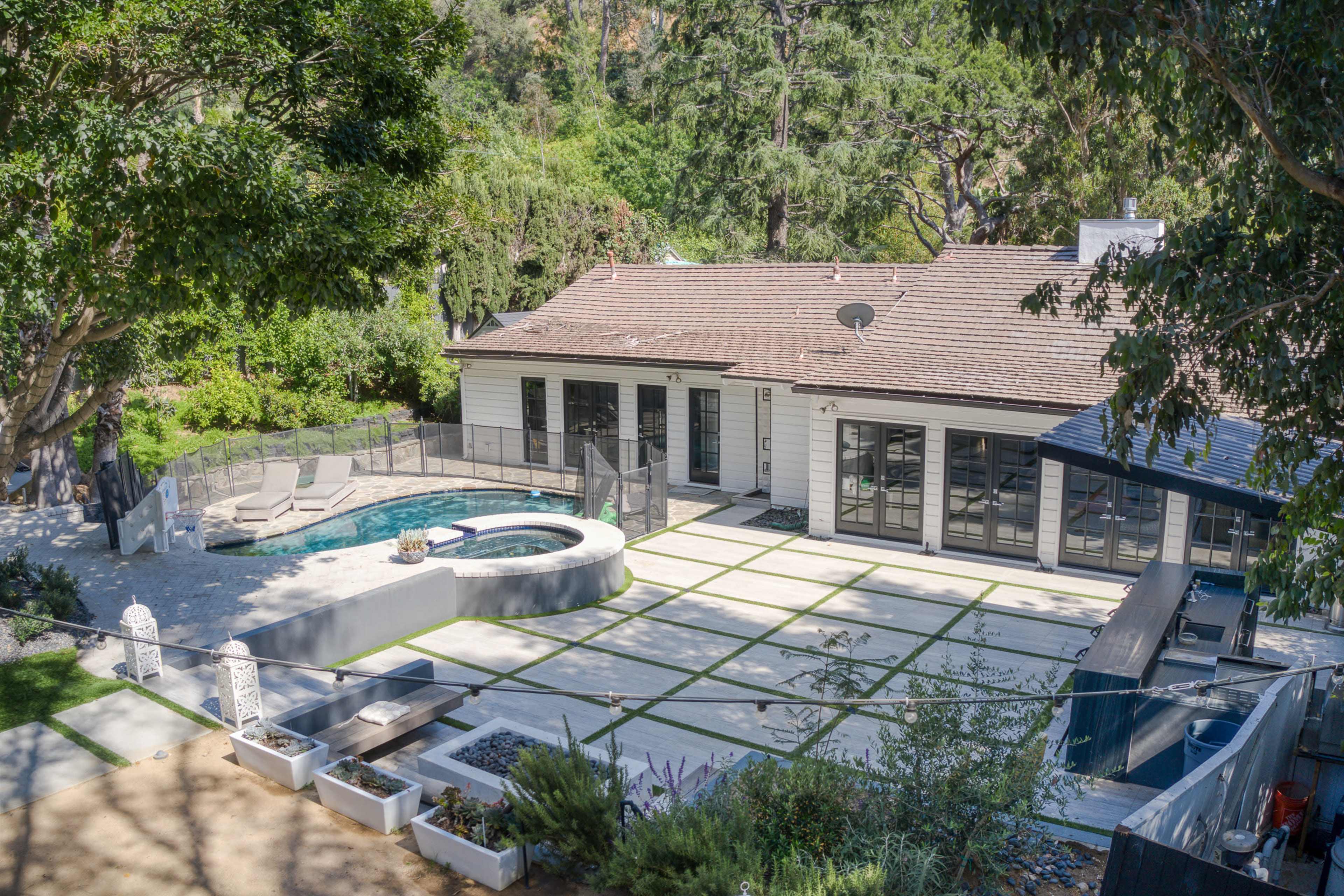 The image shows a modern single-story house with a landscaped backyard featuring a spa, seating area, and pathway surrounded by greenery.