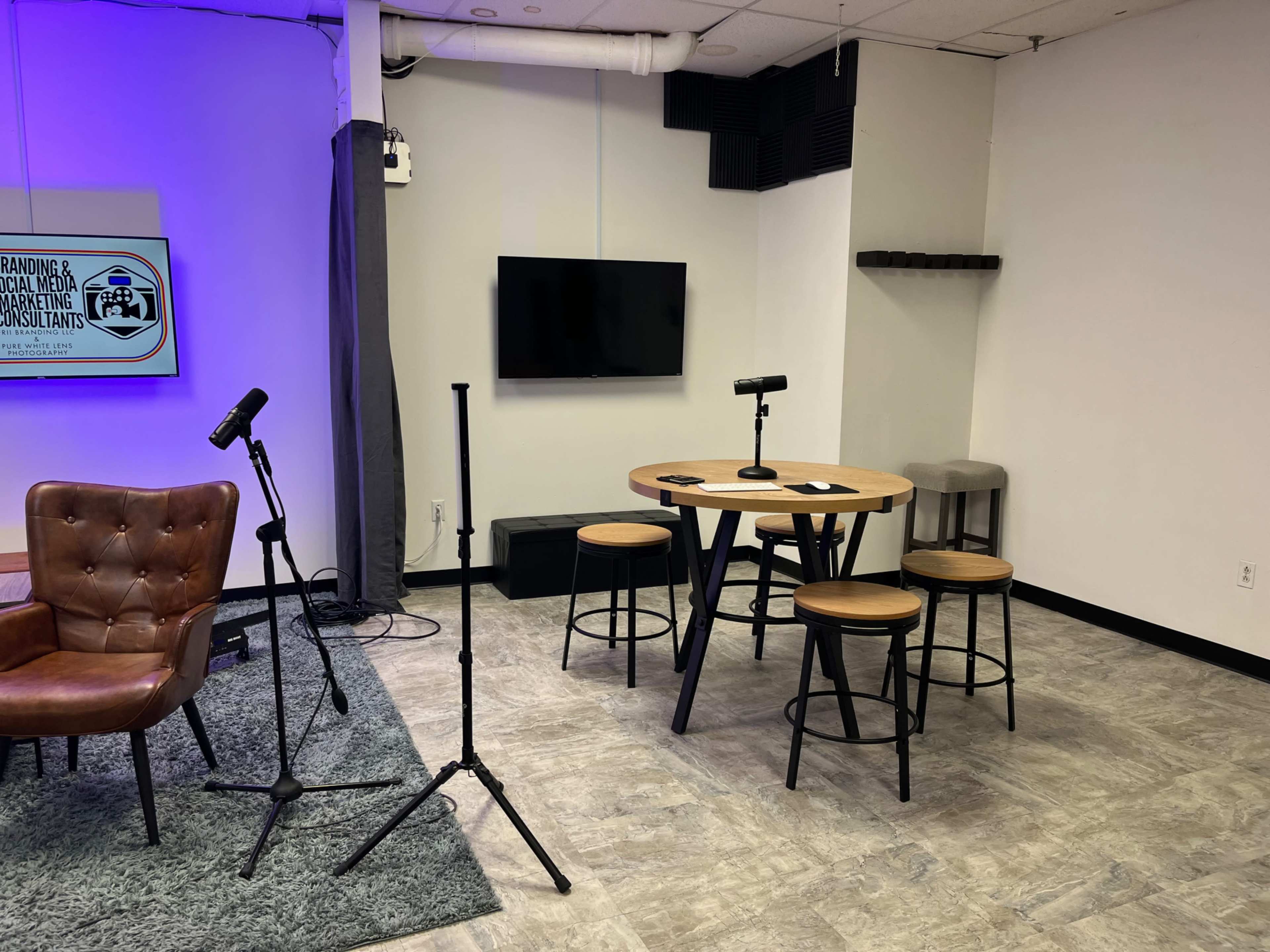 A modern studio setup featuring a circular table with four stools, three microphones on stands, a wall-mounted television, and a gray rug on the floor.