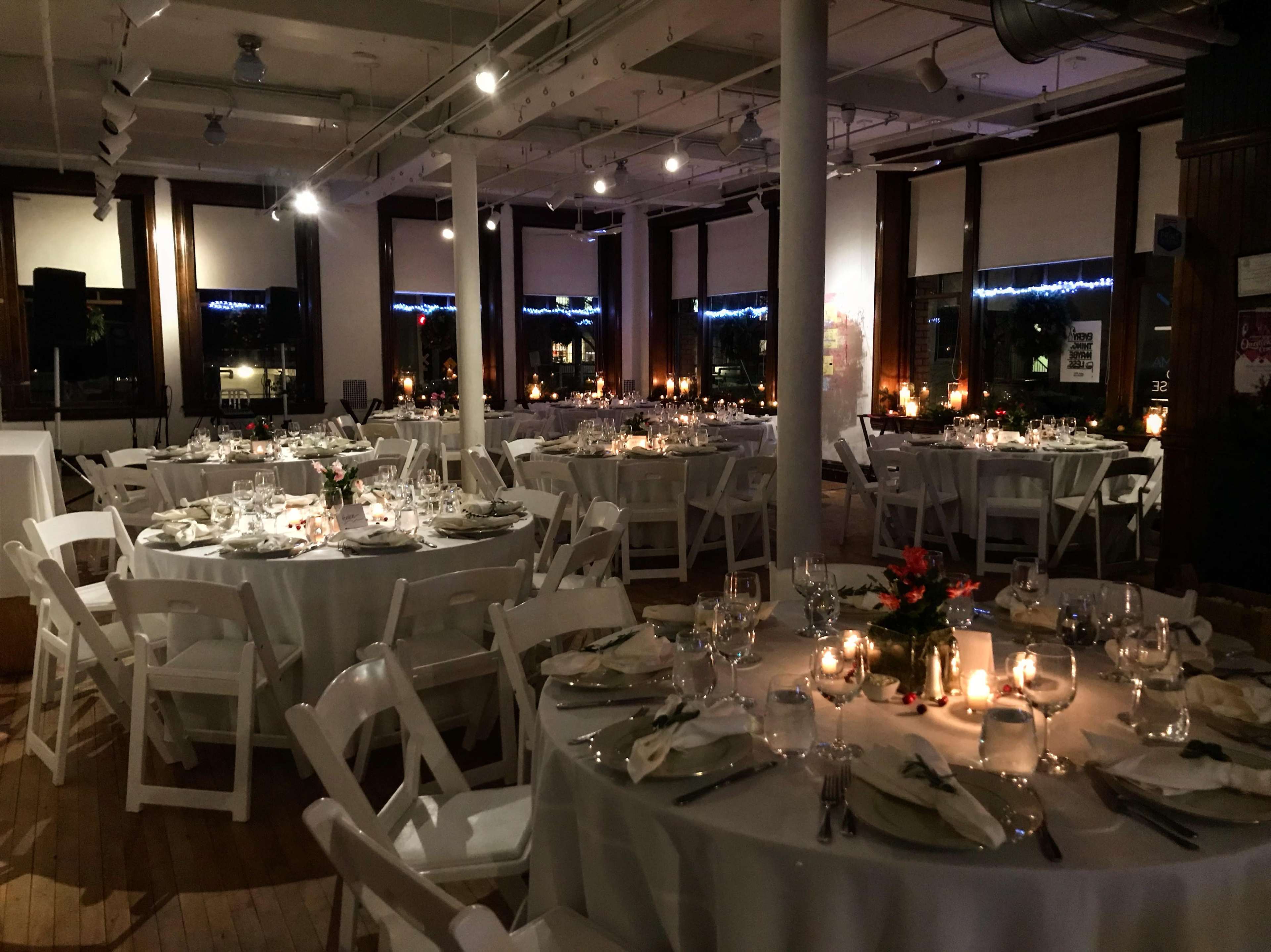 An elegantly arranged dining area with round tables set for a formal event, featuring white tablecloths, glassware, and centerpieces, illuminated by candlelight.