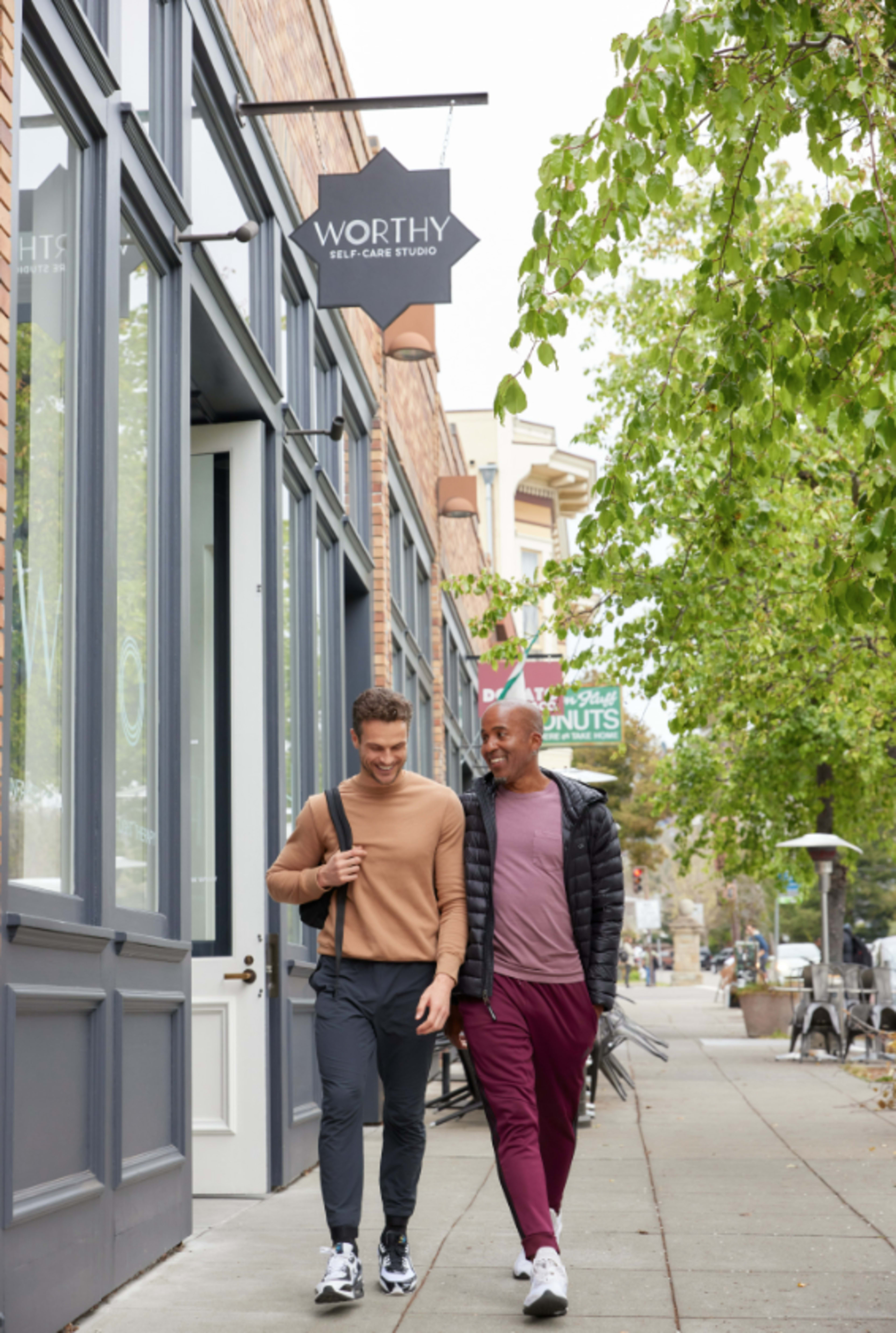 Two men walk along a sidewalk in front of a storefront labeled "Worthy Self-Care Studio."