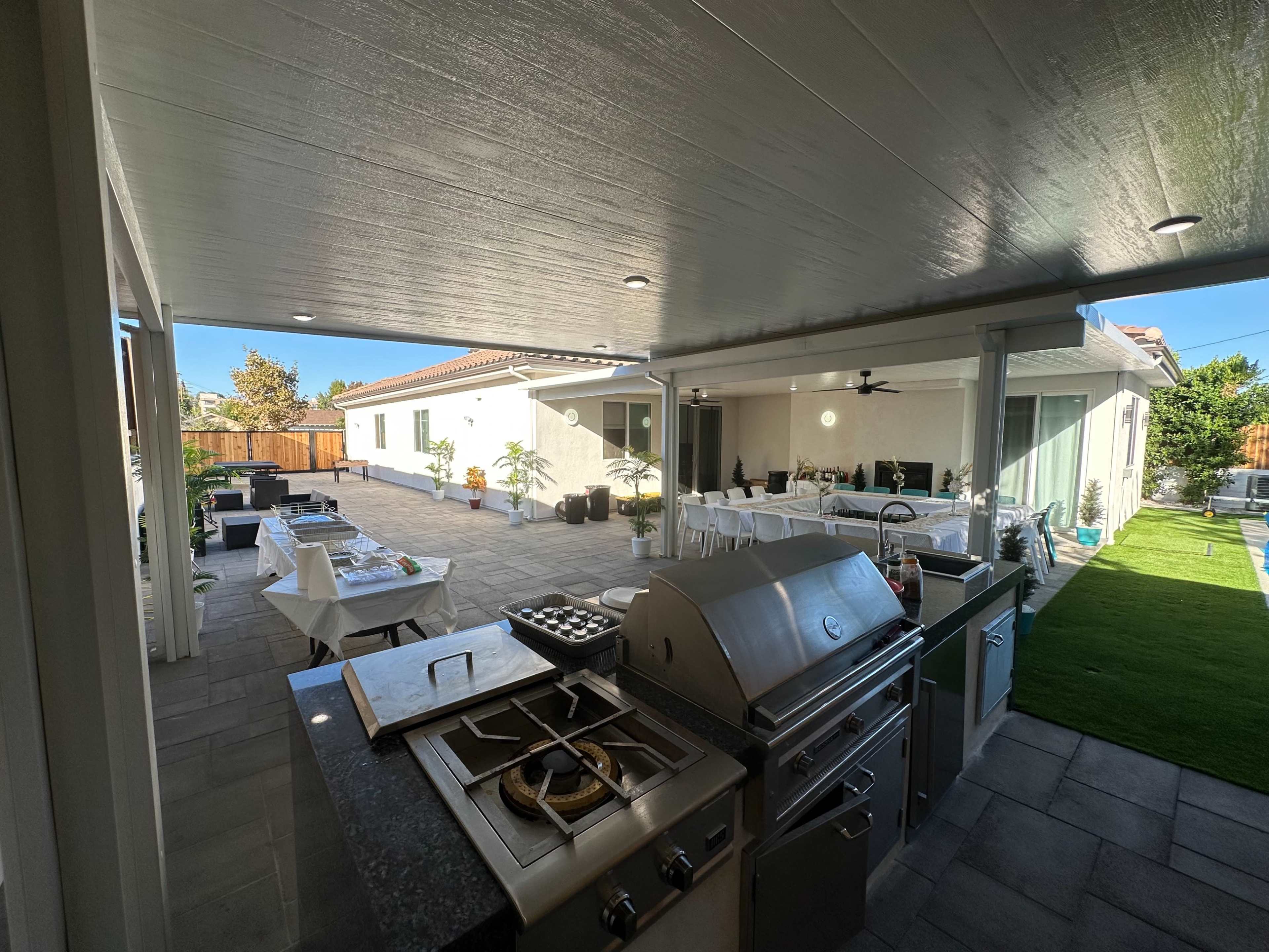 An outdoor patio area with a built-in grill and seating arrangements, featuring a lawn and dining setup under a covered structure.