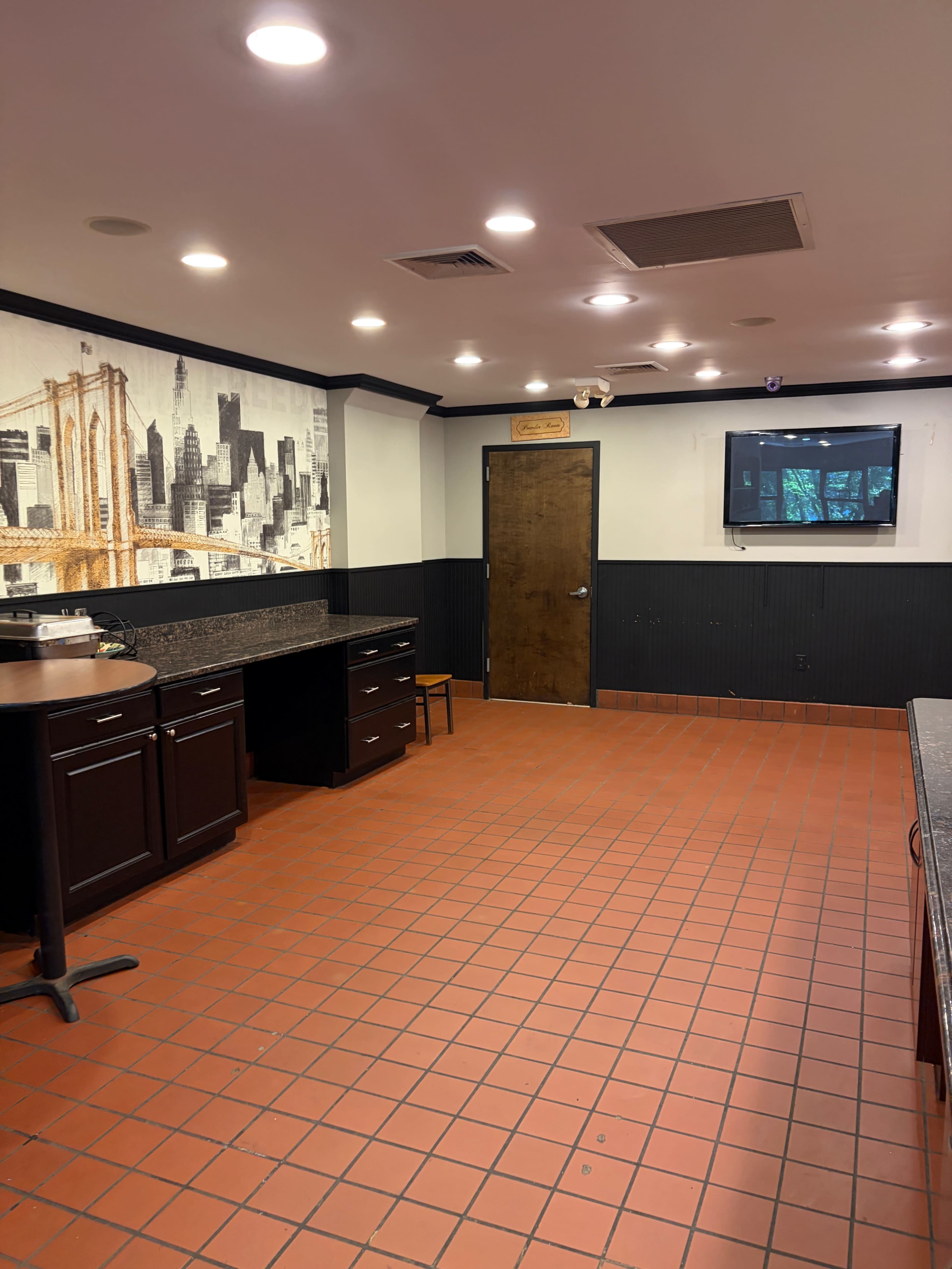 The image shows a kitchen area with red tiled flooring, dark cabinets, a small chair, a television mounted on the wall, and a mural of a city skyline.