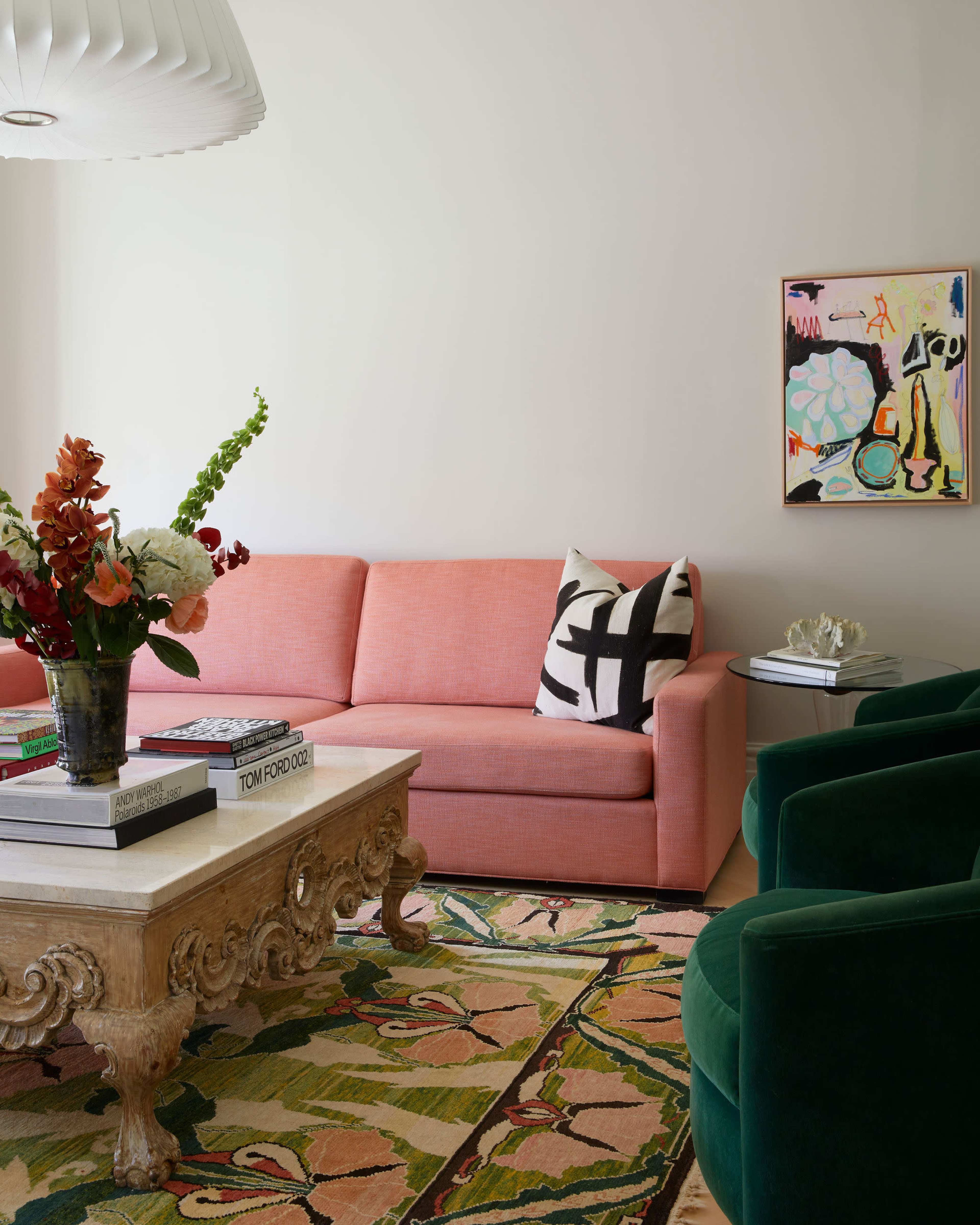 The image depicts a living room featuring a pink couch, a decorative coffee table, a colorful rug, and two green upholstered chairs, complemented by floral arrangements and a vibrant piece of artwork on the wall.