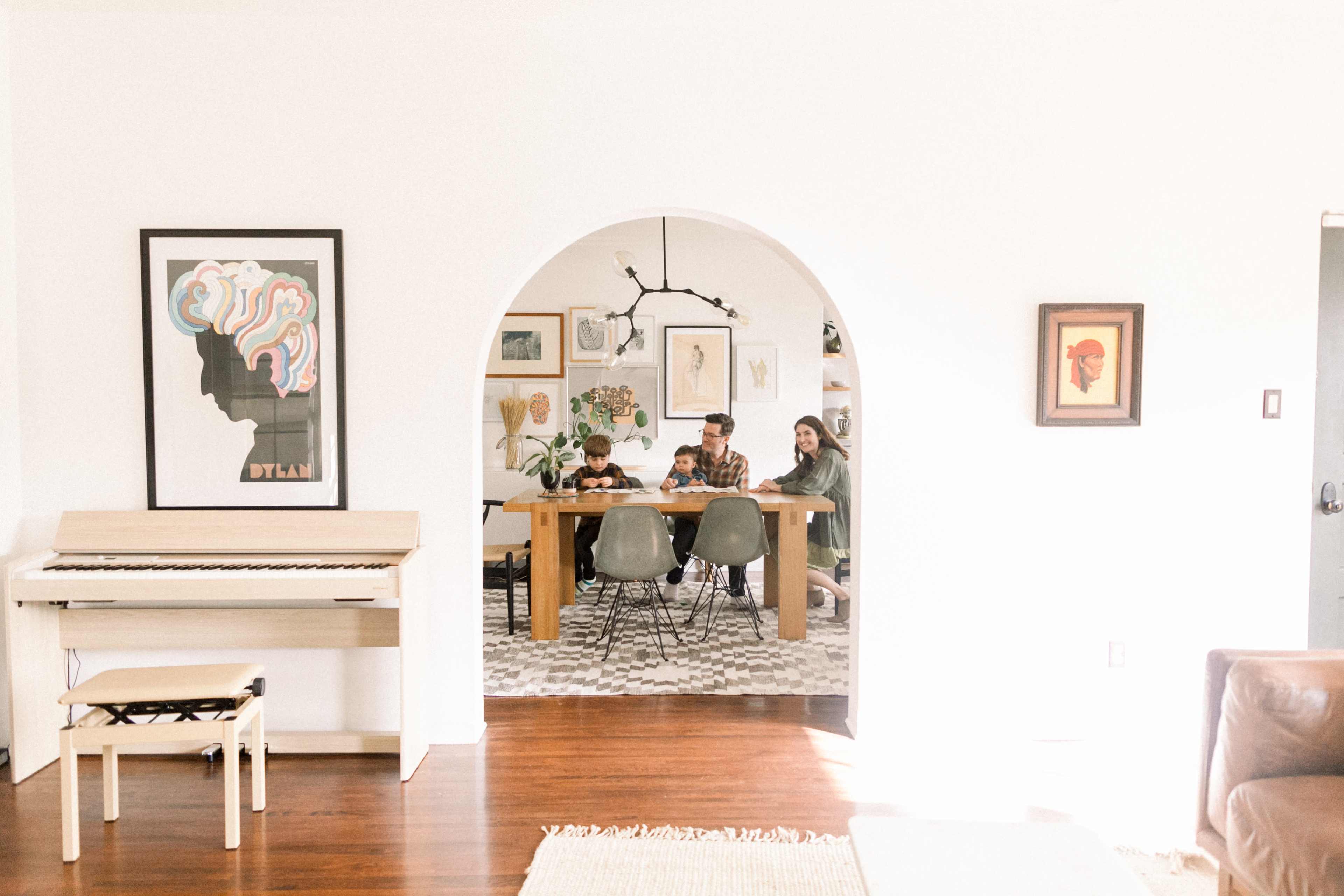 A family sits around a dining table in a bright room, with a piano and framed artwork visible in the background.