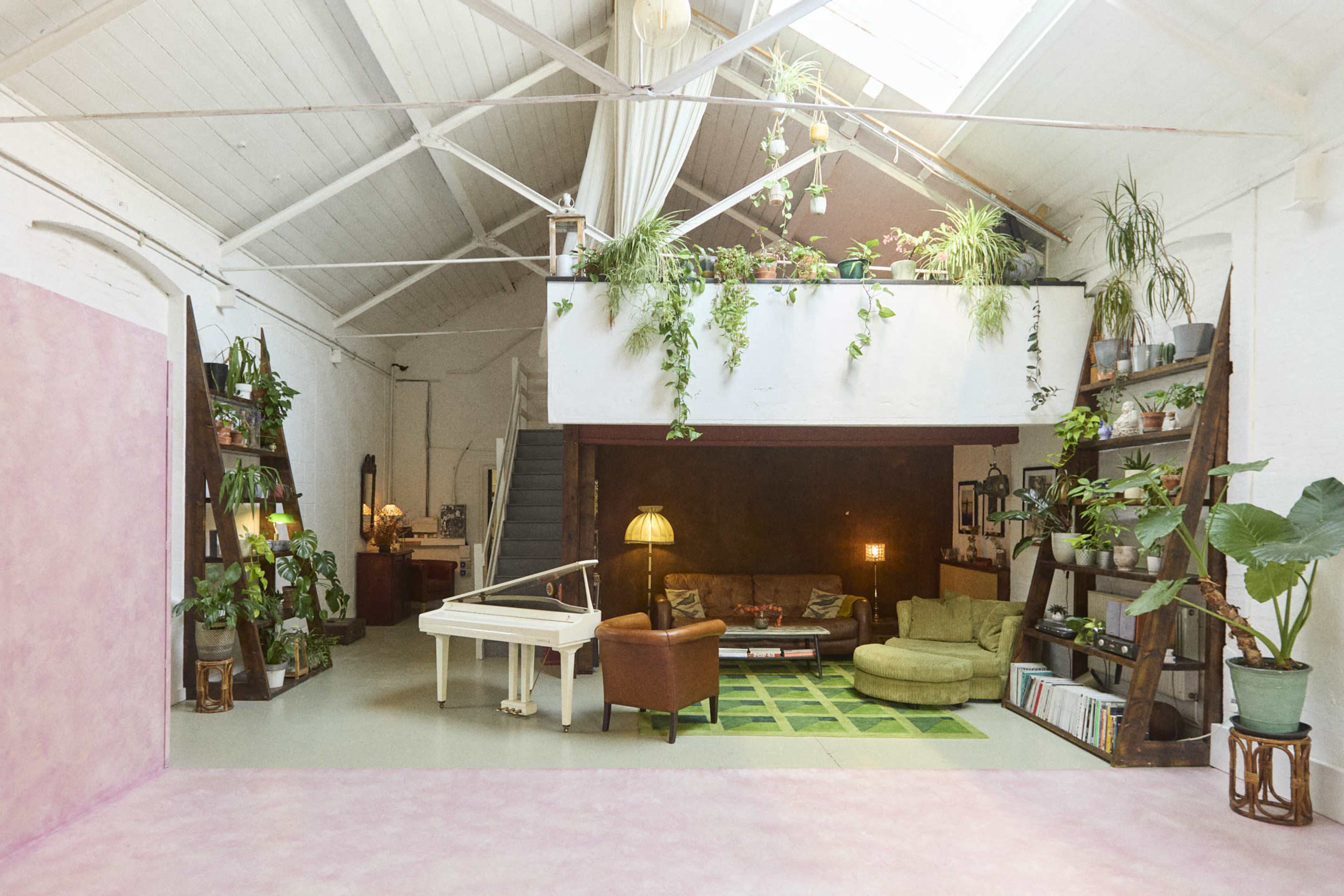 A spacious, brightly lit loft with green plants, a white piano, a green sofa, and shelves filled with books and decorative items.