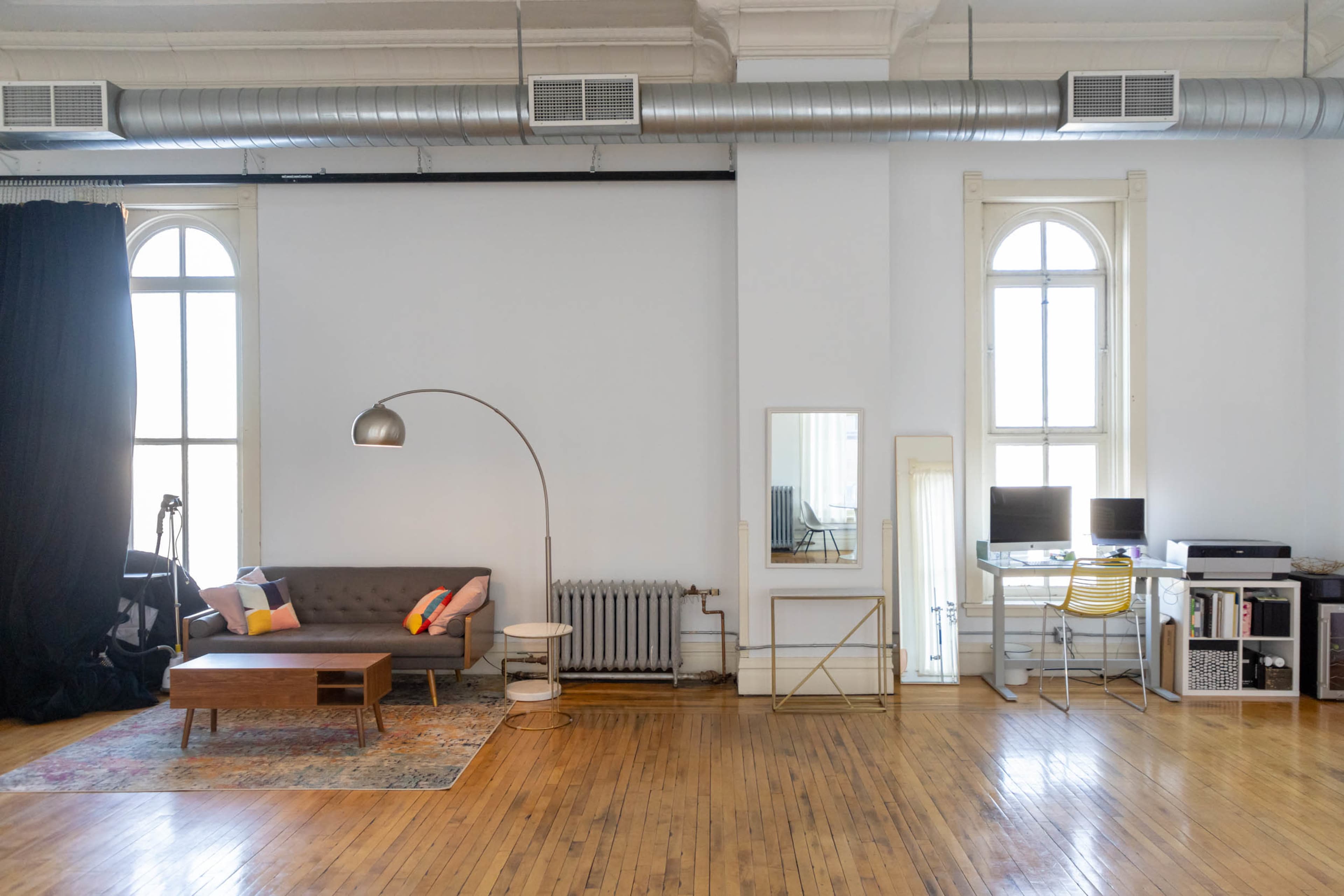 The image shows a spacious living area with a gray sofa, wooden coffee table, and desk setup, featuring large windows and wooden flooring.