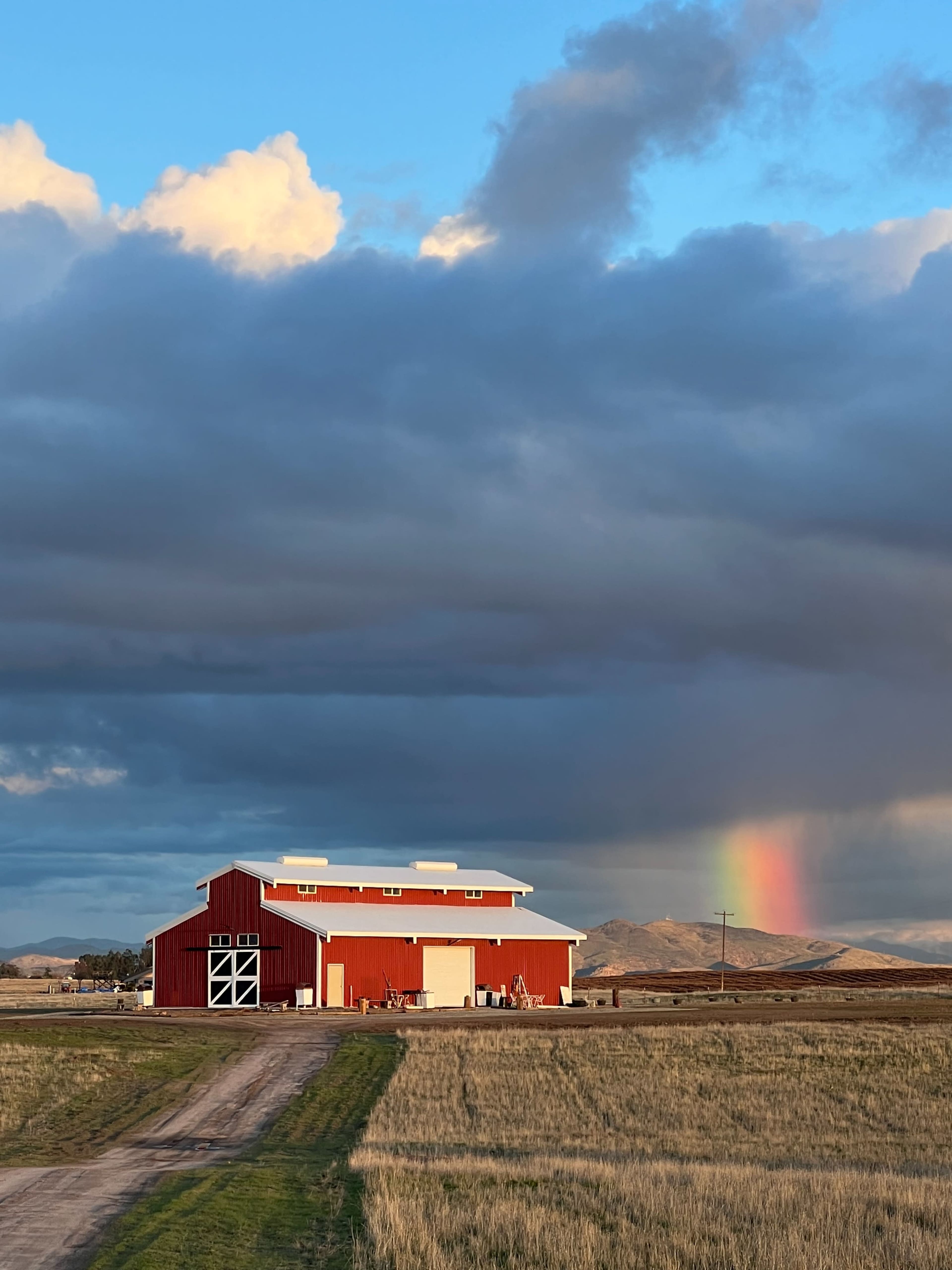 A red barn stands in a field under a cloudy sky, with a rainbow appearing in the distance.
