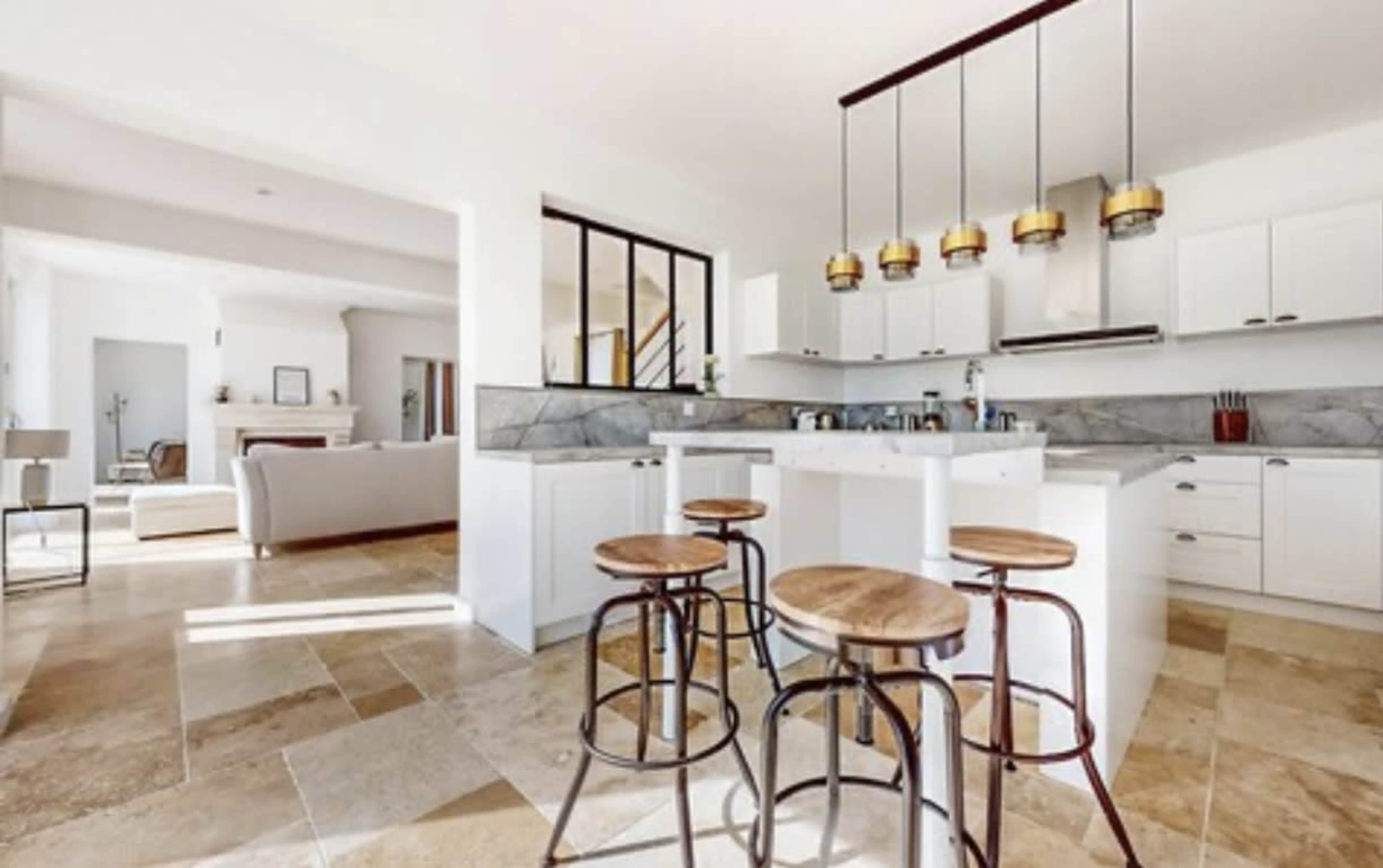 The image shows a modern kitchen with white cabinets, a marble countertop, and a breakfast bar featuring wooden stools.