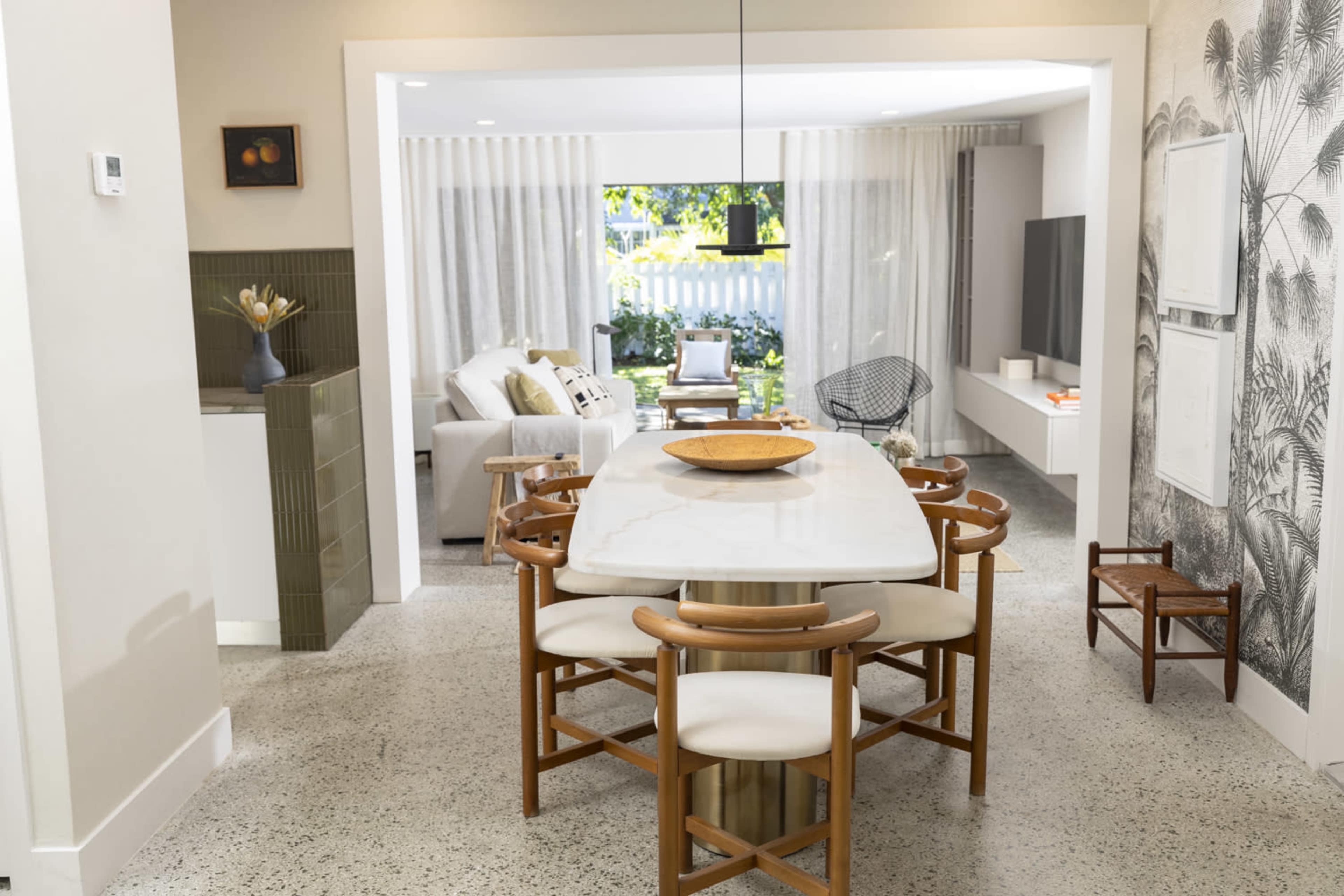 A dining area features a large white table surrounded by wooden chairs, leading to a living space with large windows and a couch.