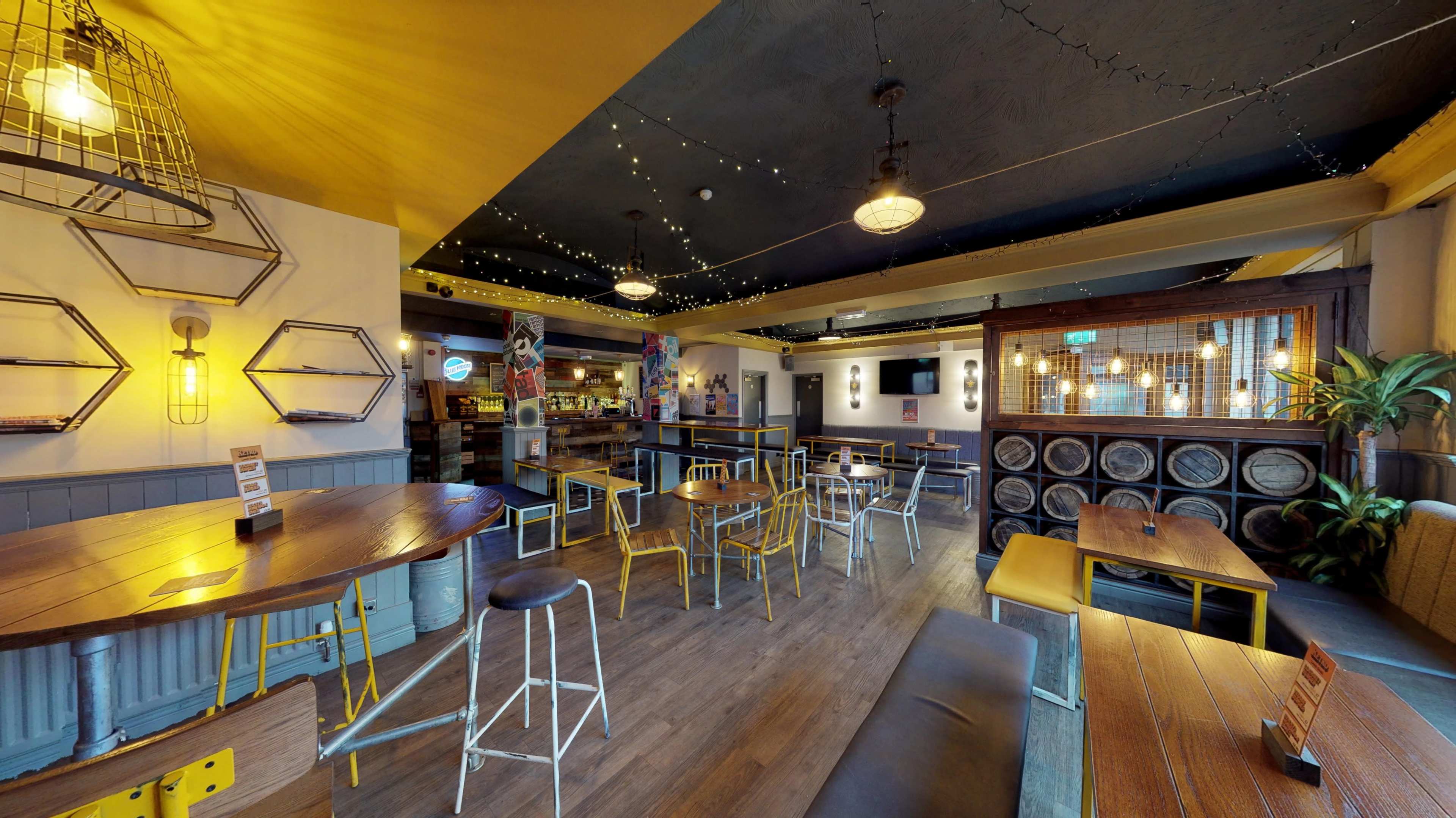 The image depicts an empty restaurant interior with wooden tables, metal chairs, and decorative lighting, featuring a bar area in the background.
