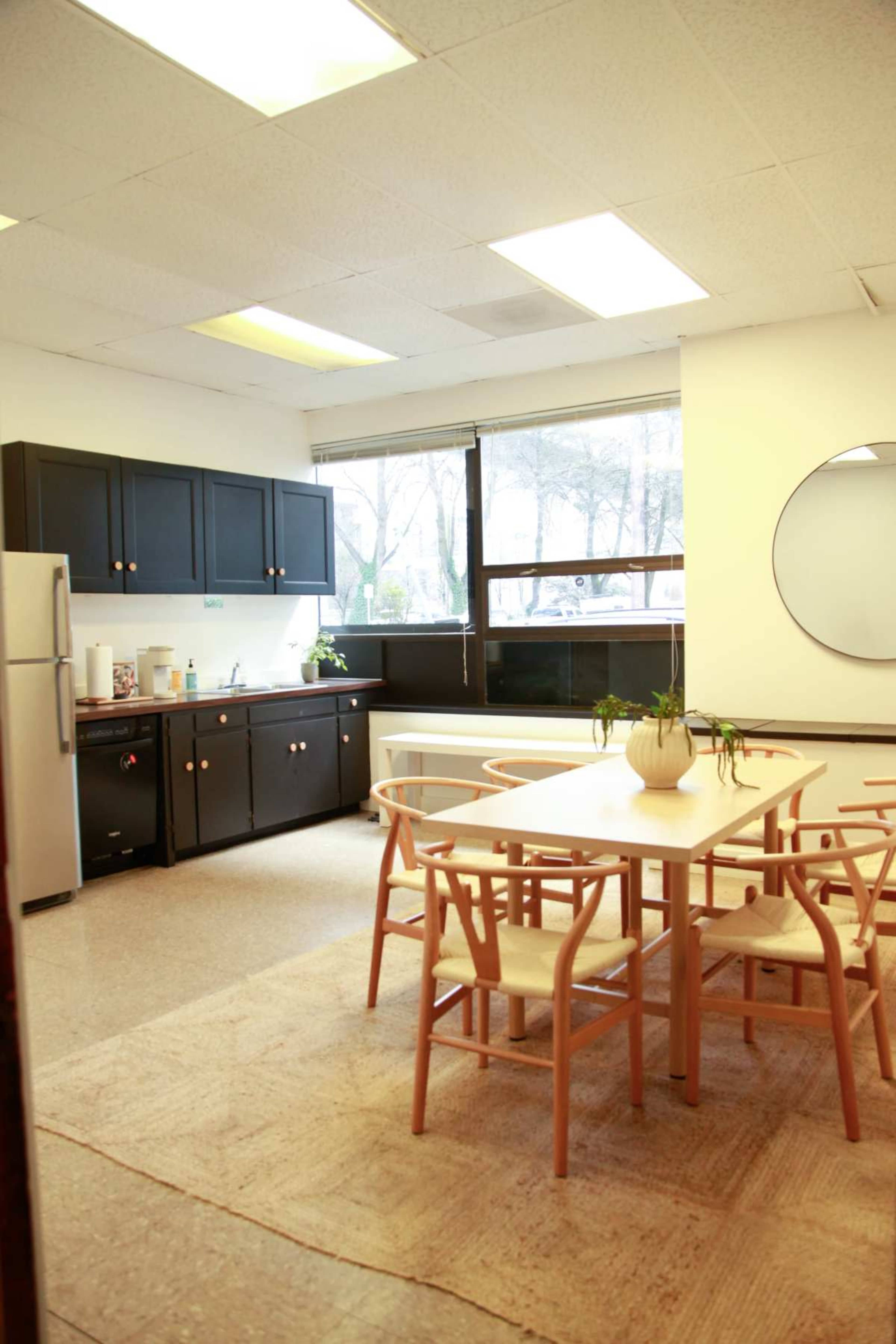 The image shows a bright kitchen and dining area with a wooden table surrounded by chairs, a refrigerator, and cabinets along the wall.