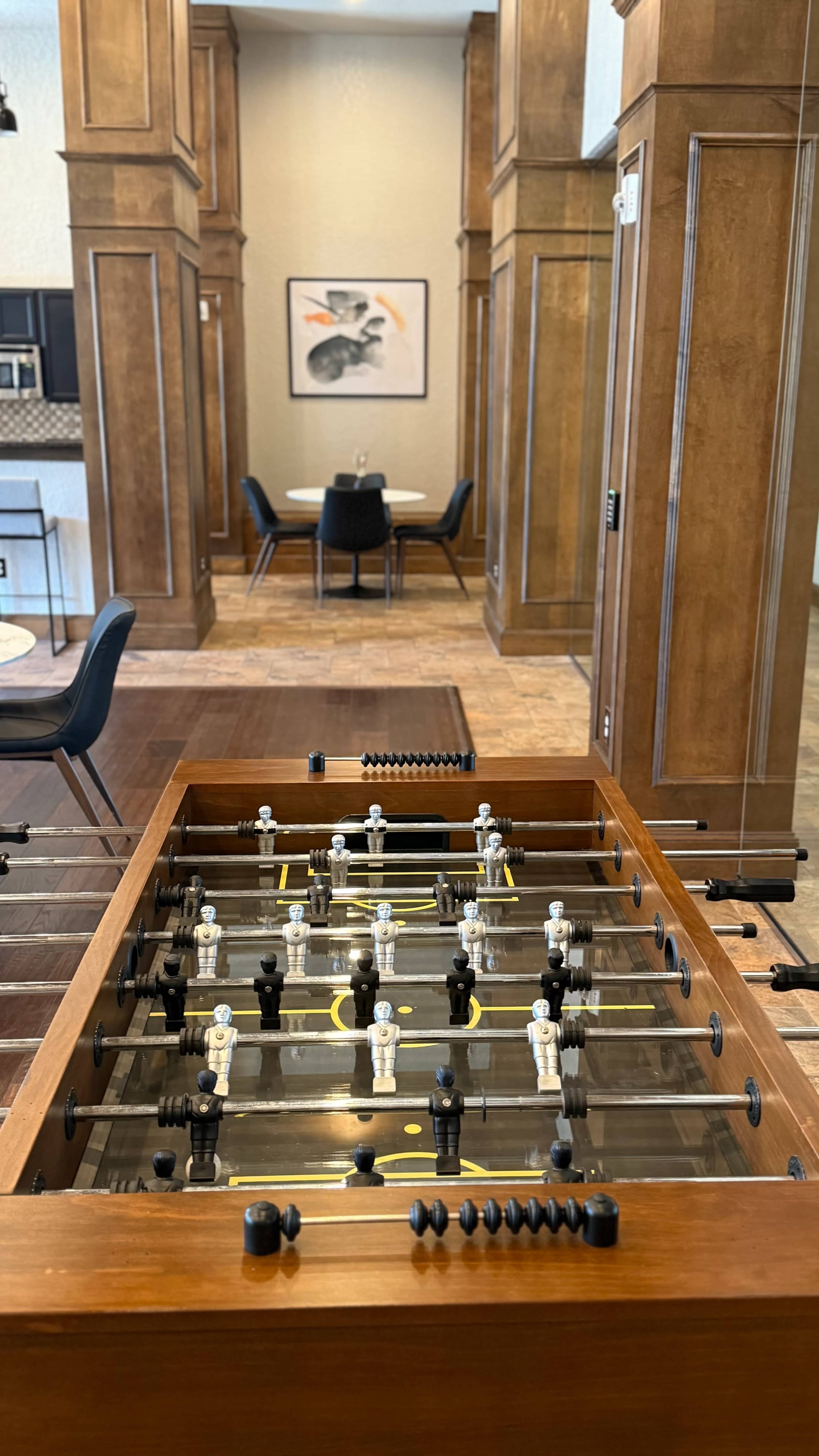 A foosball table is positioned in the foreground, with a dining area visible in the background featuring a table and chairs.