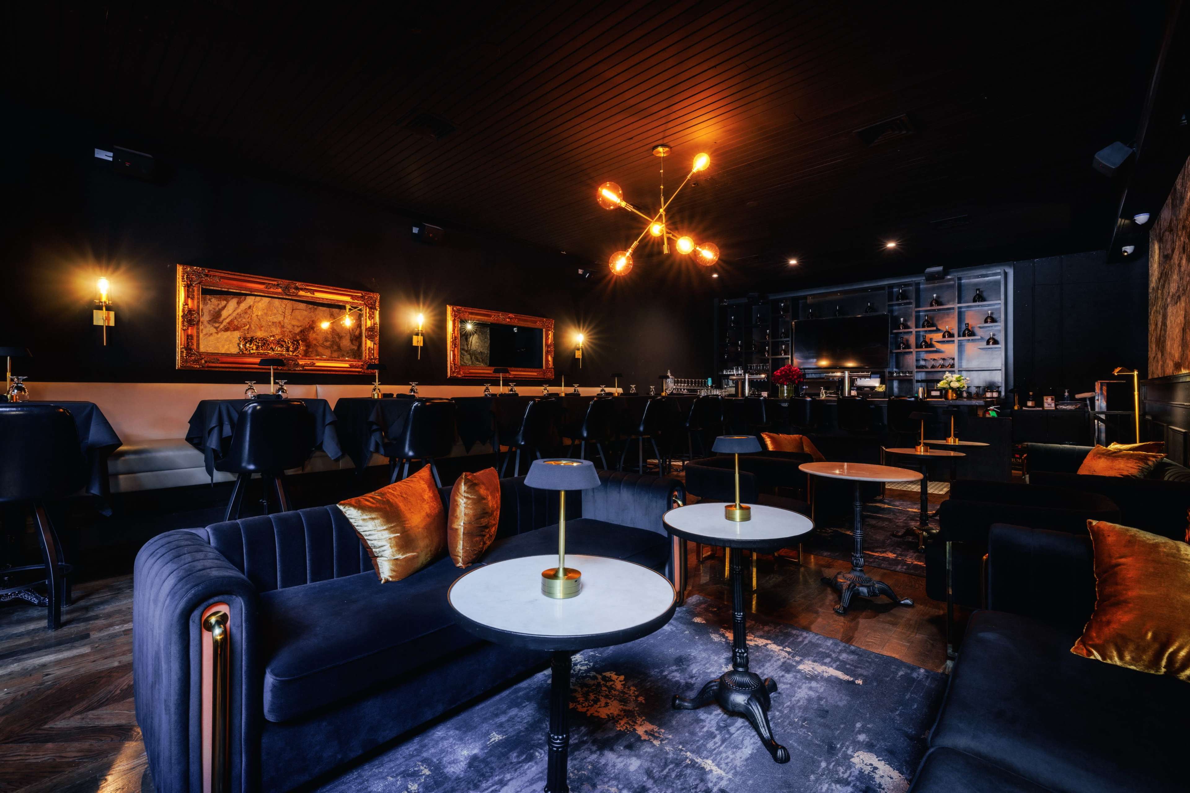 The image shows a sleek, modern lounge with plush seating, polished tables, and a stylish bar area illuminated by decorative light fixtures.