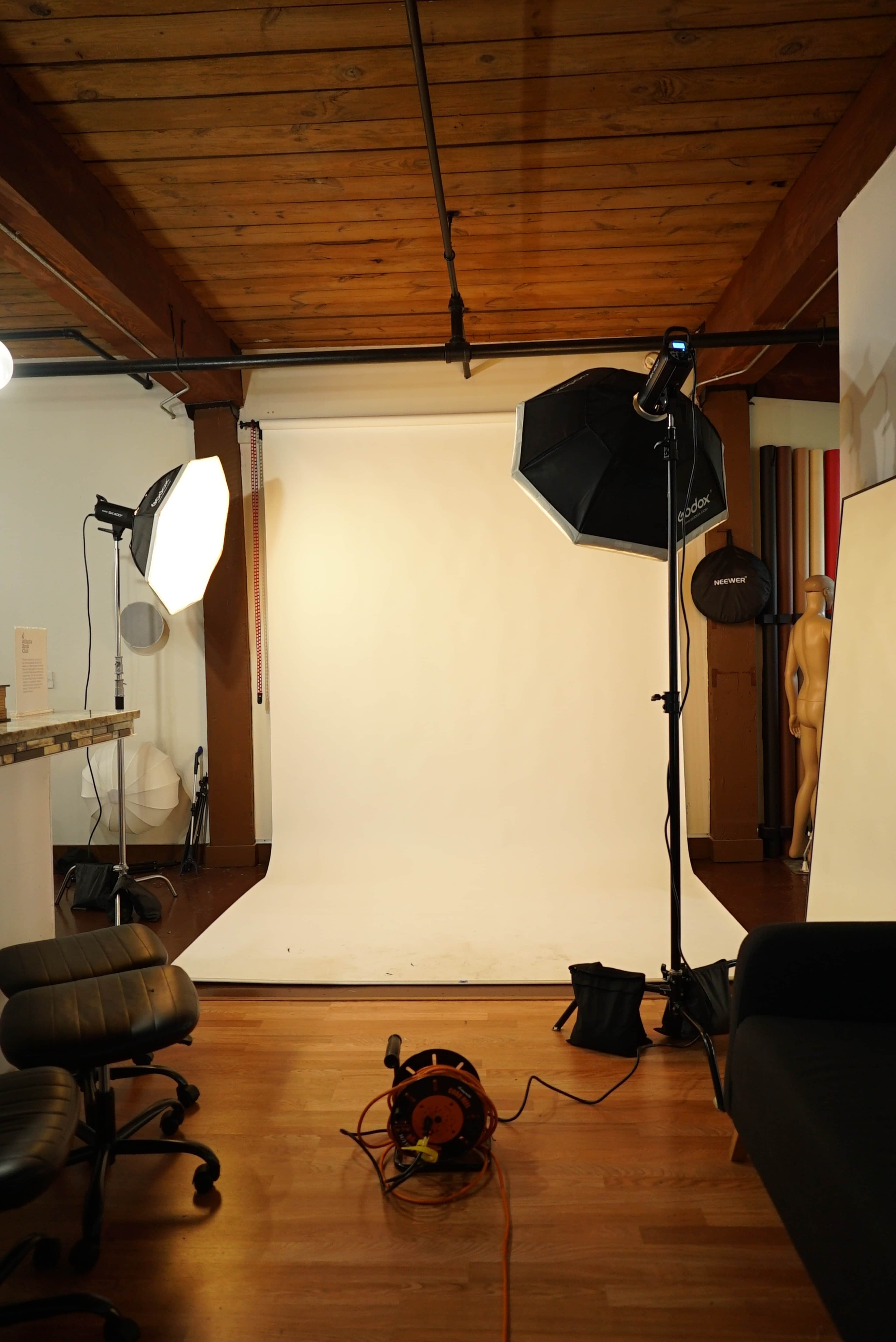 The image shows a photography studio with a seamless white backdrop, lighting equipment, and a few chairs in the foreground.