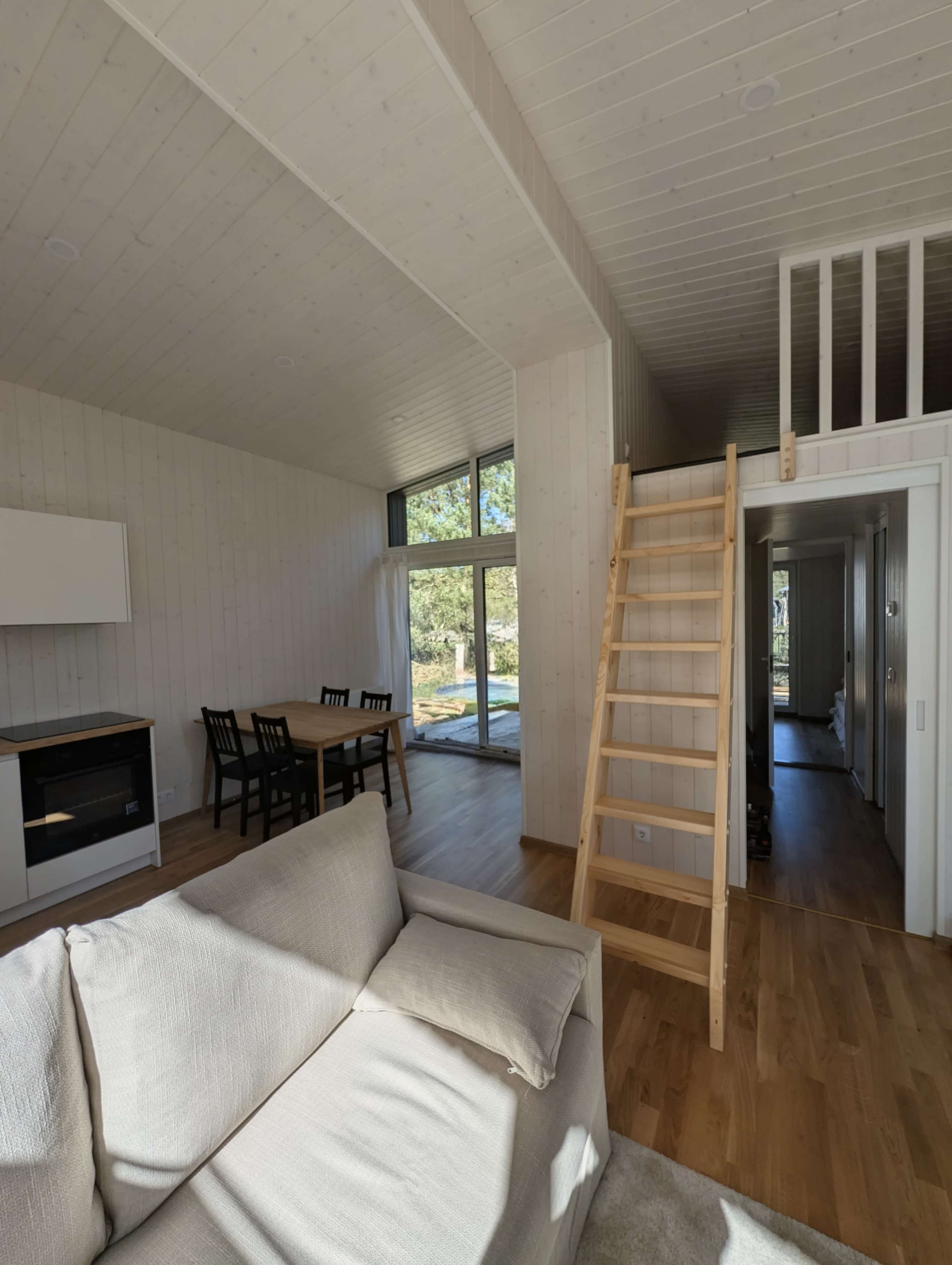 The image shows a modern, minimalist living space with a wooden staircase leading to a loft area, and a dining table set in a bright, open room.