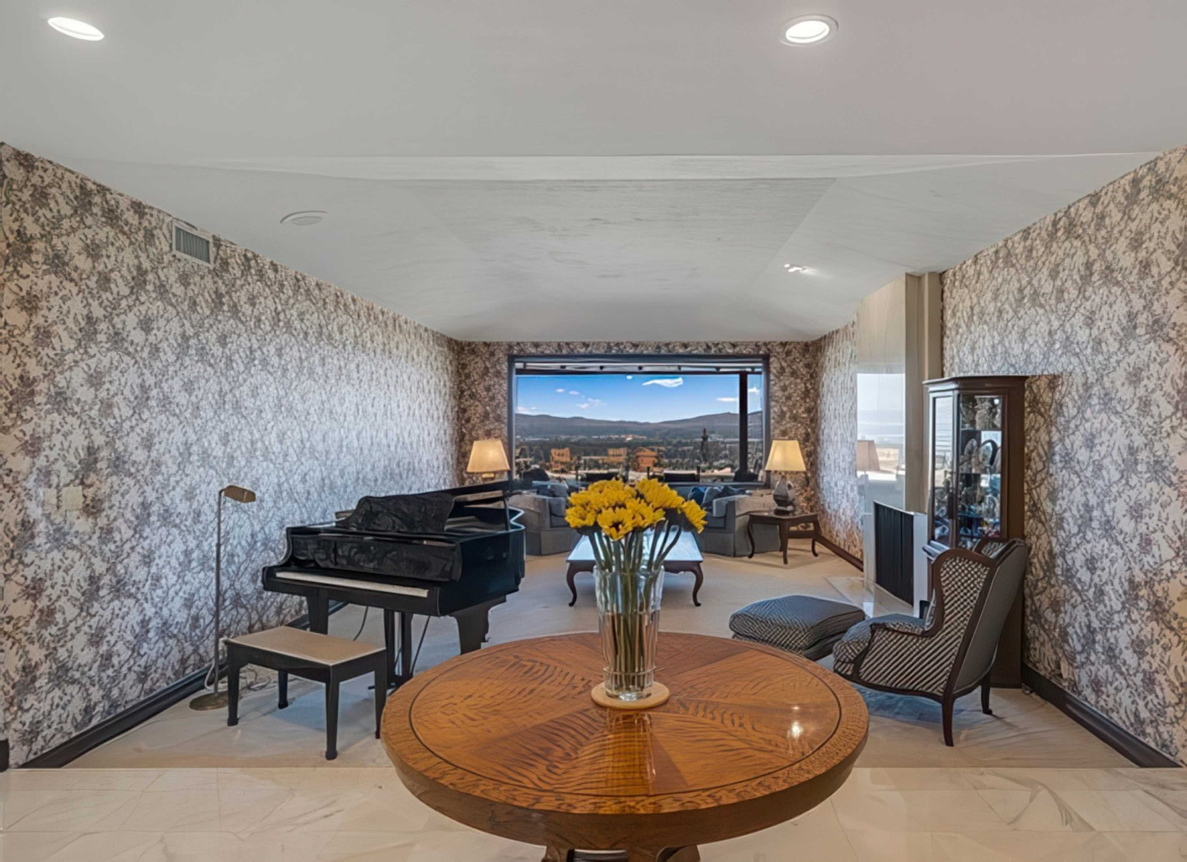 The image shows a living room featuring a grand piano, a round wooden table with a vase of yellow flowers, and large windows with a mountain view.