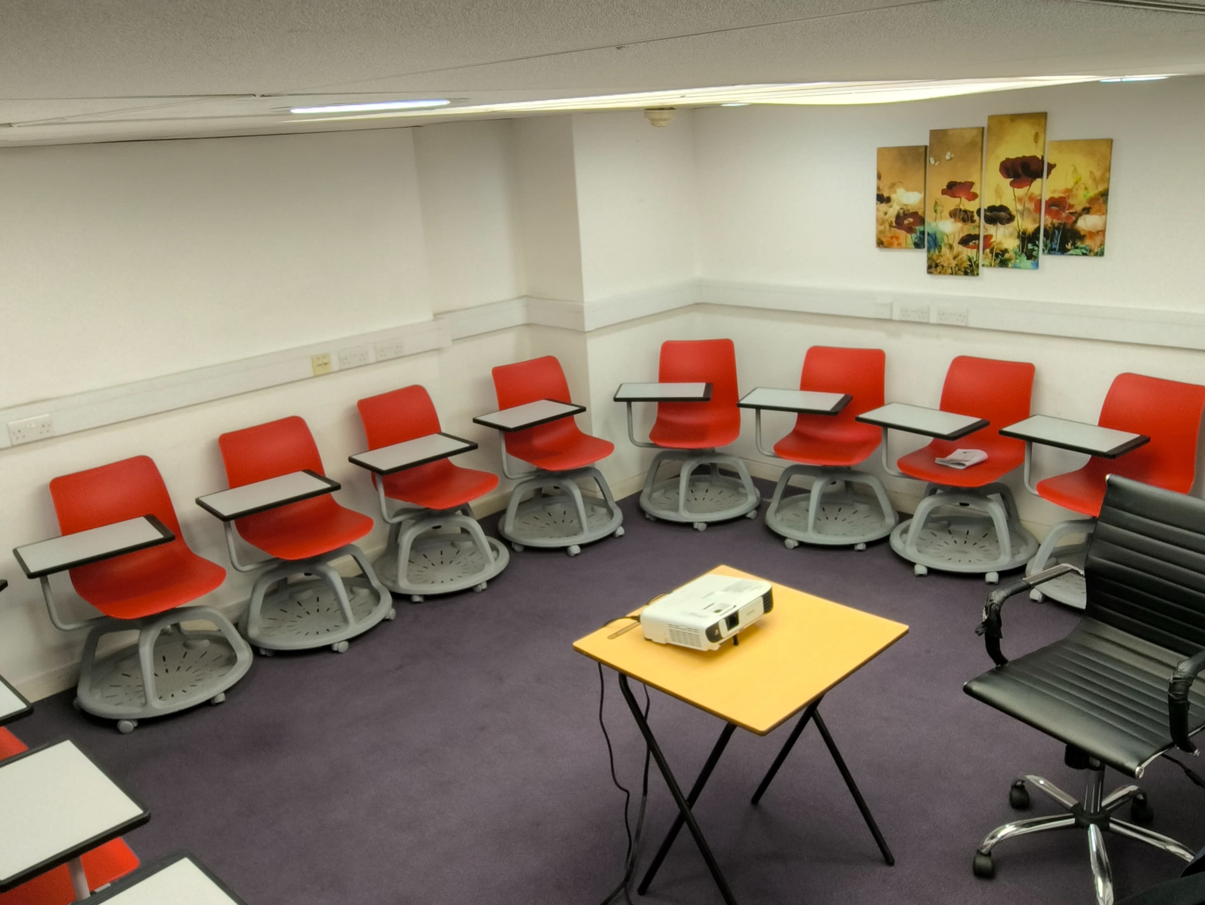 The image shows a small meeting room with red chairs arranged in a circular formation around a central table, accompanied by a projector and paintings on the wall.