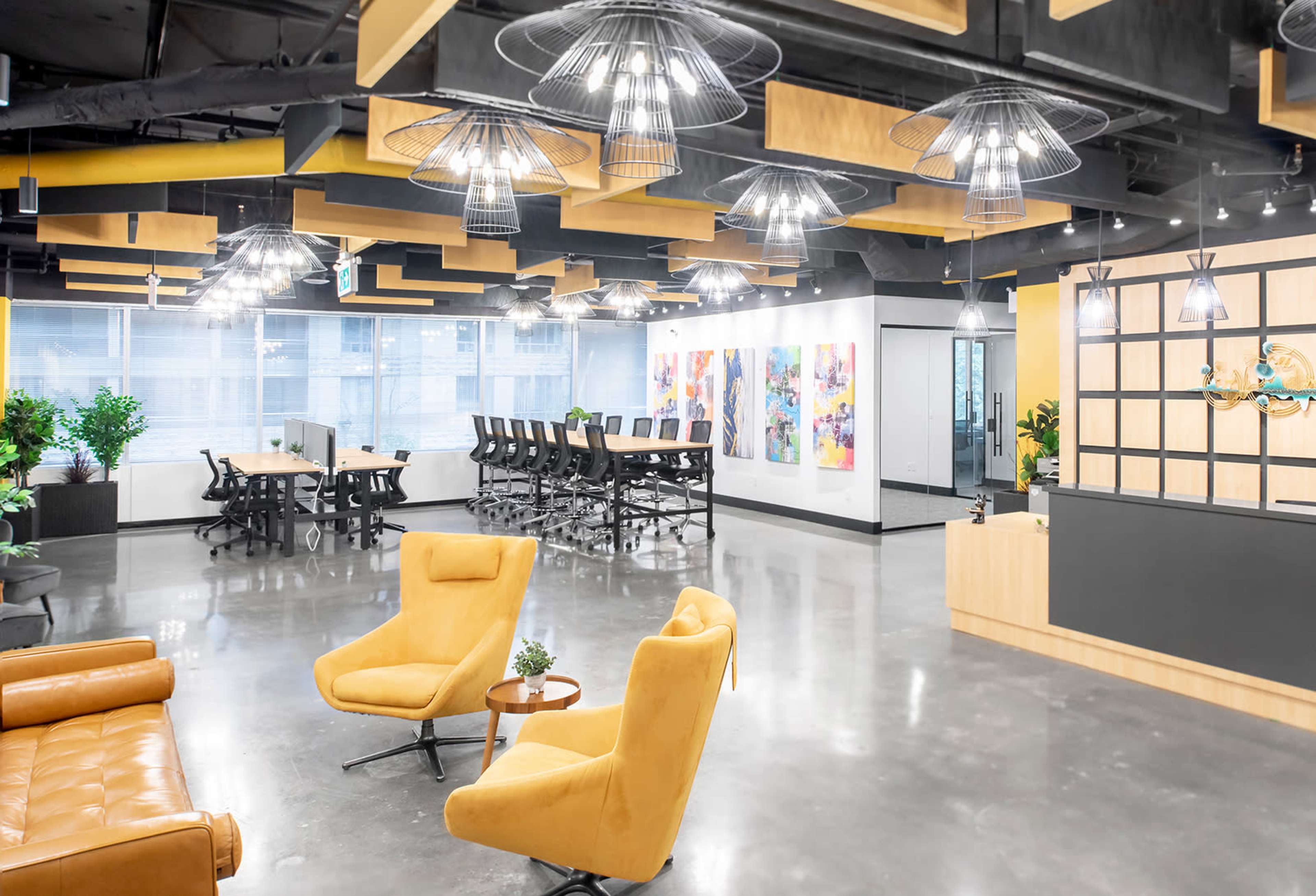 The image shows a modern office space featuring yellow armchairs, a small table, and a large conference table surrounded by ergonomic chairs, with artistic wall decor and ample natural light.