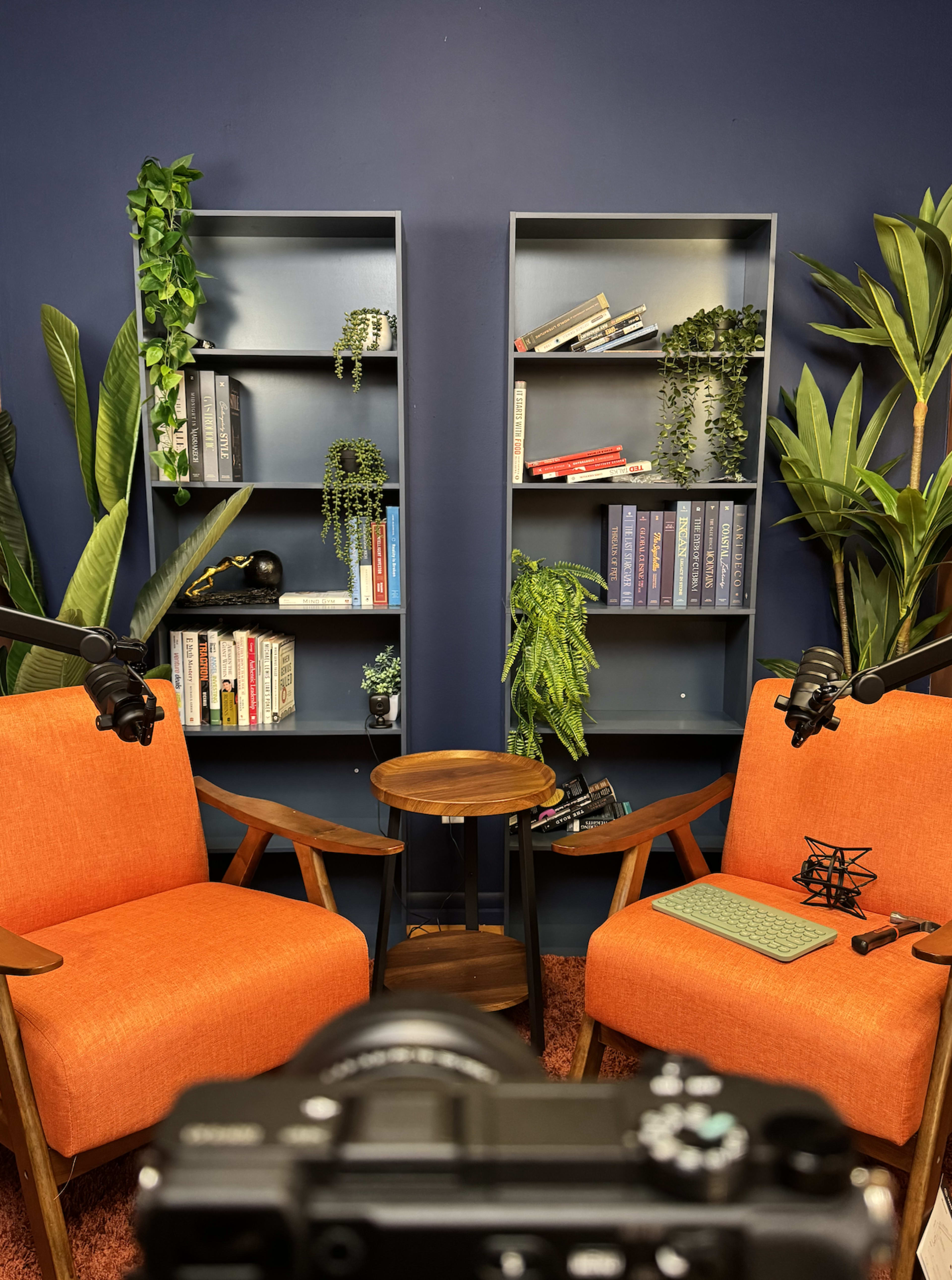 A camera is pointed at a cozy conversation setup featuring two orange chairs, a small table, and shelves filled with books and plants against a blue wall.