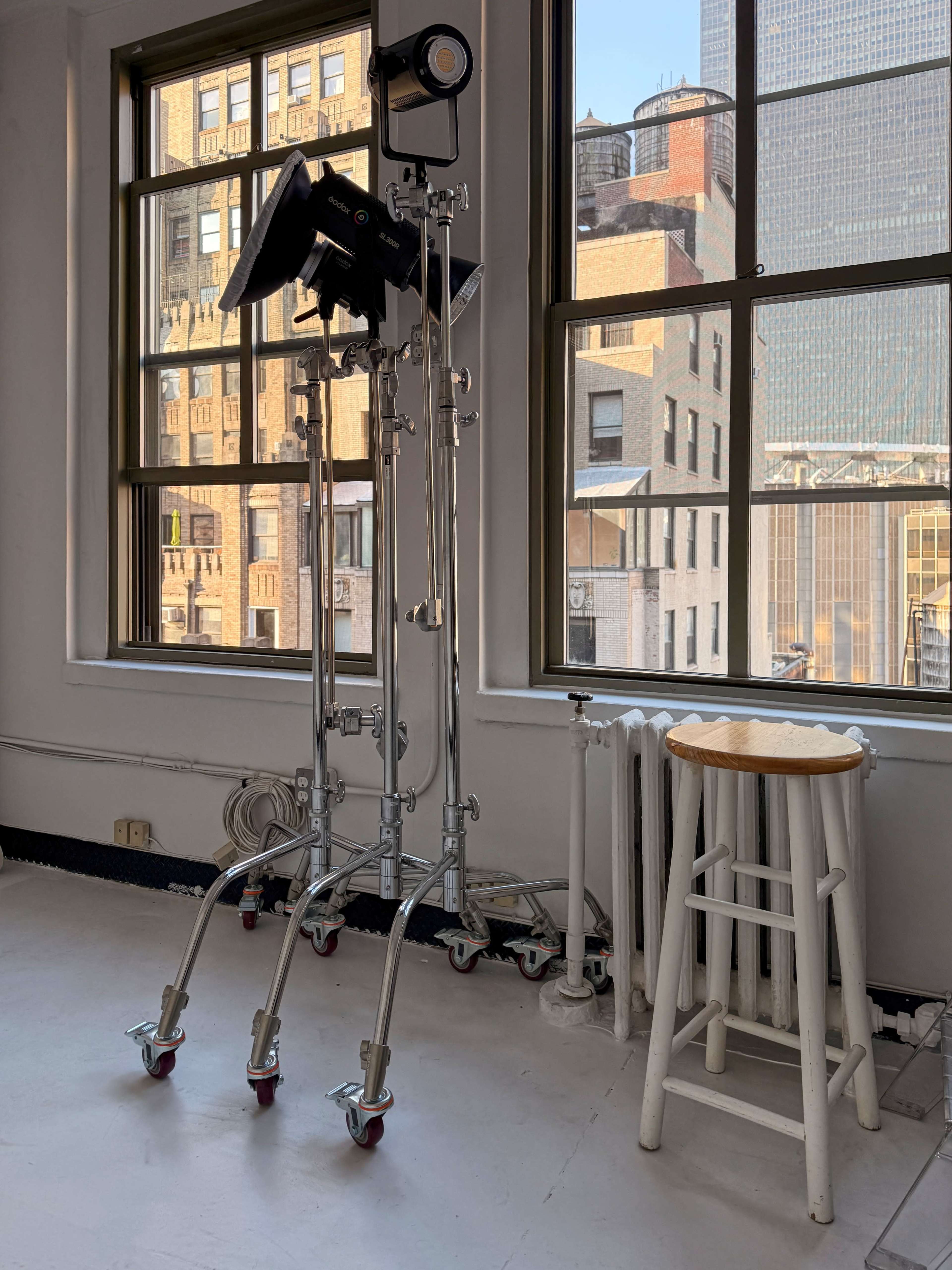 The image shows a photography setup featuring a tall light stand with a softbox and a white bar stool positioned near large windows overlooking a cityscape.
