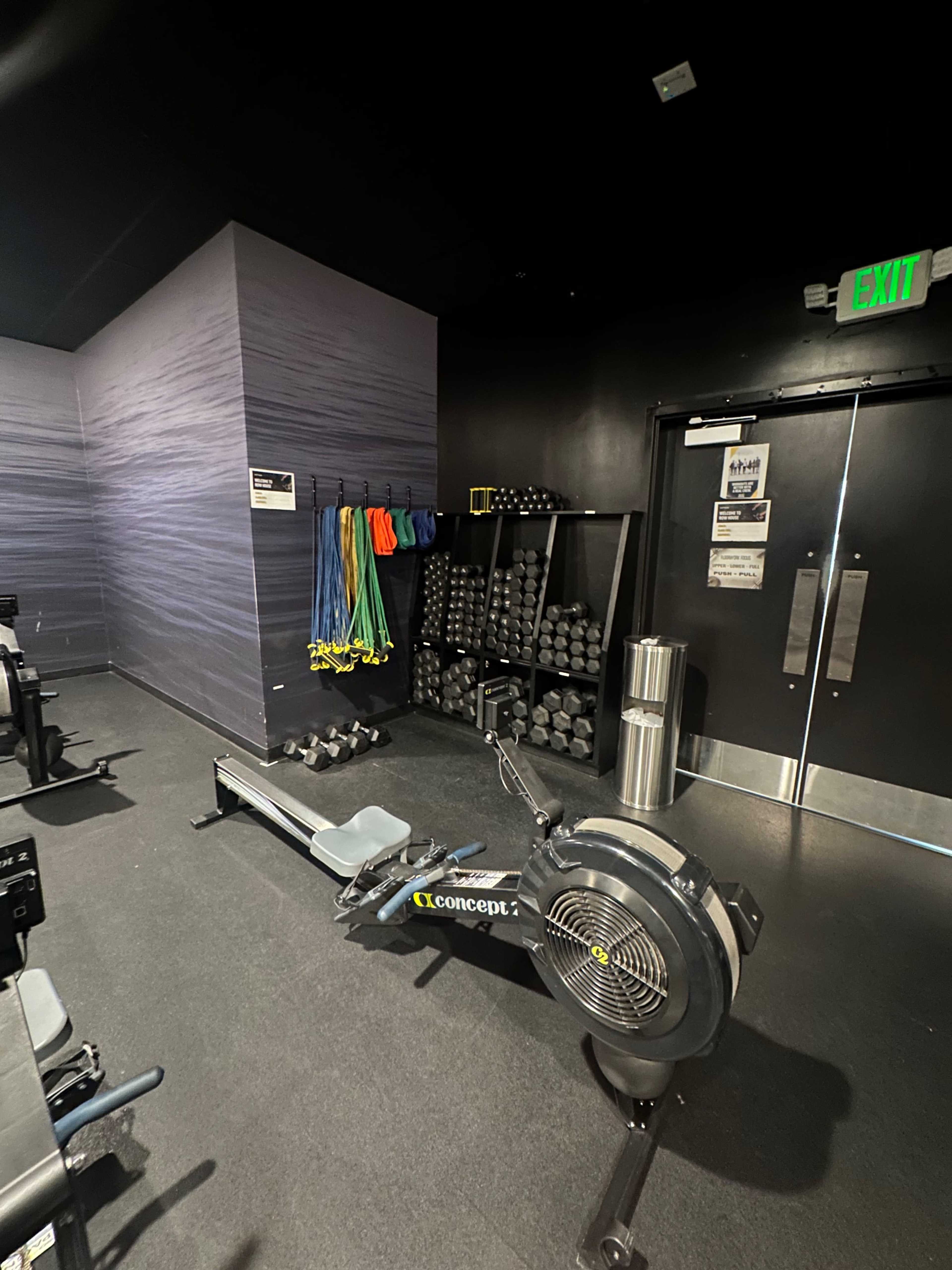 The image shows a gym area with a rowing machine, free weights, and resistance bands against a wall.