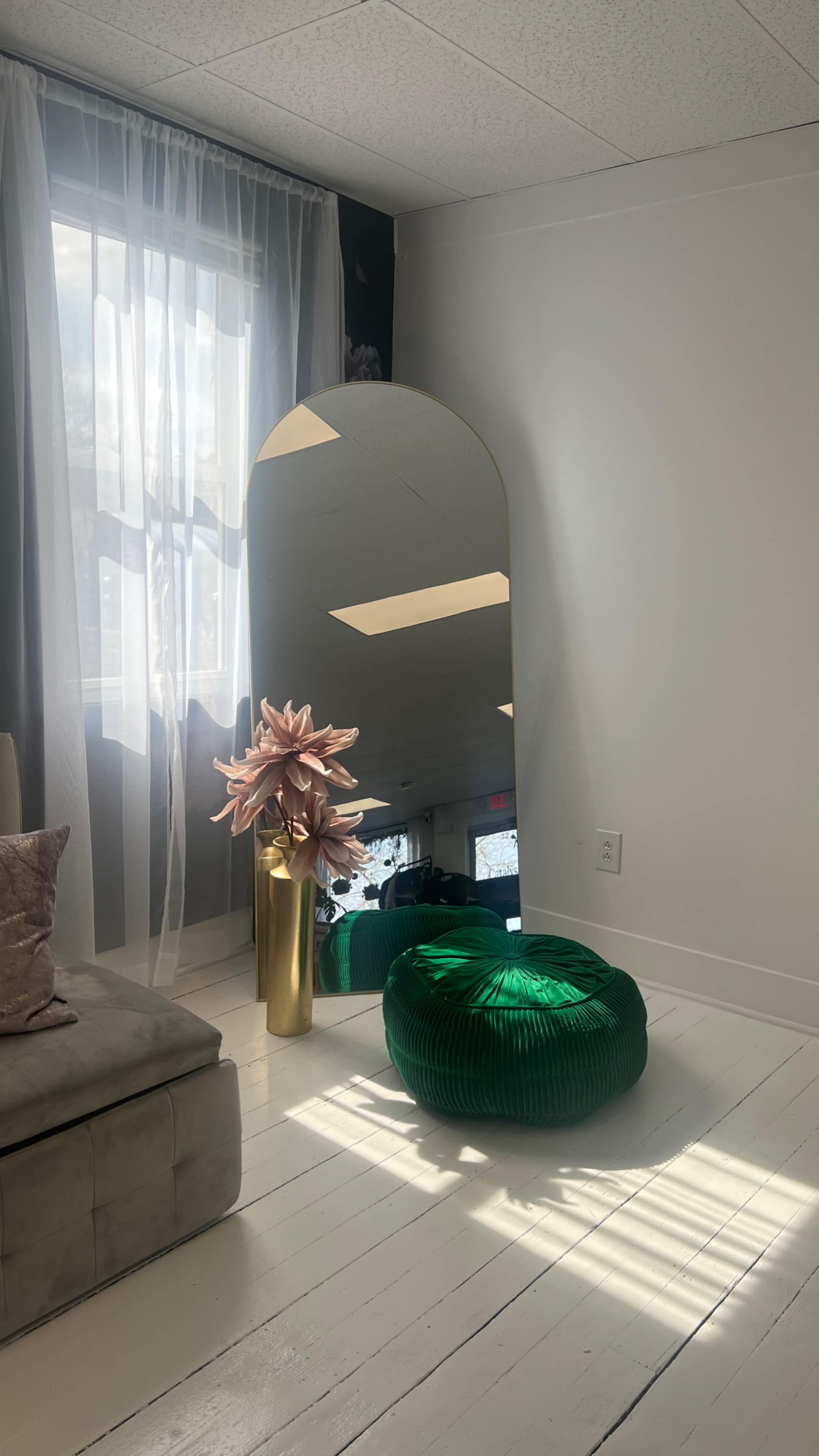 The image shows a bright room featuring a large mirror, a green pouf, a tall vase with a pink flower arrangement, and a light-colored sofa.